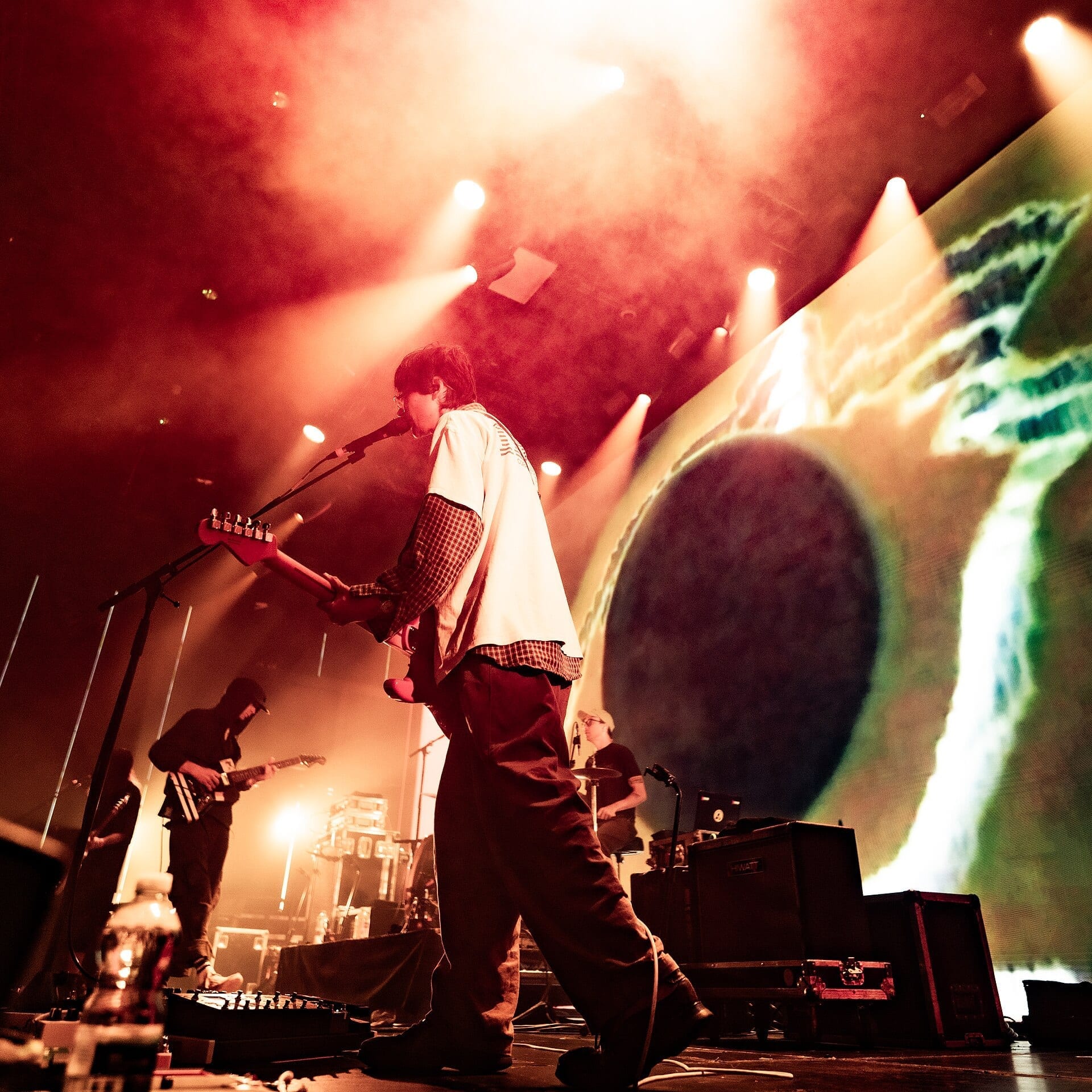 A DIIV guitarist sings into a microphone on a red-lit stage, with bandmates performing behind him in front of a large colorful video backdrop.