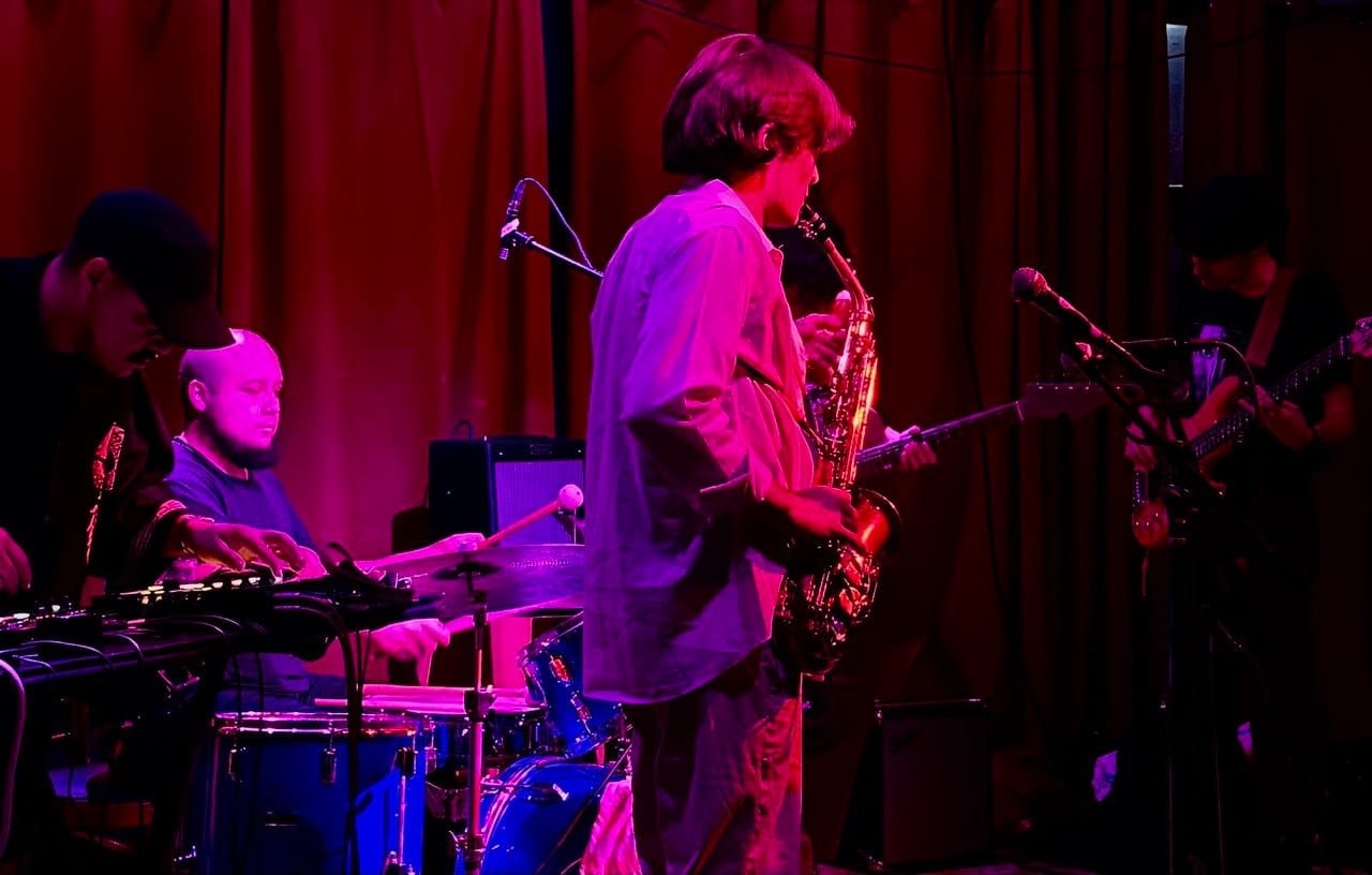Five musicians play a small venue stage under pink light in front of a red curtain, a saxophonist at the center, with drums, keys and a guitarist alongside.