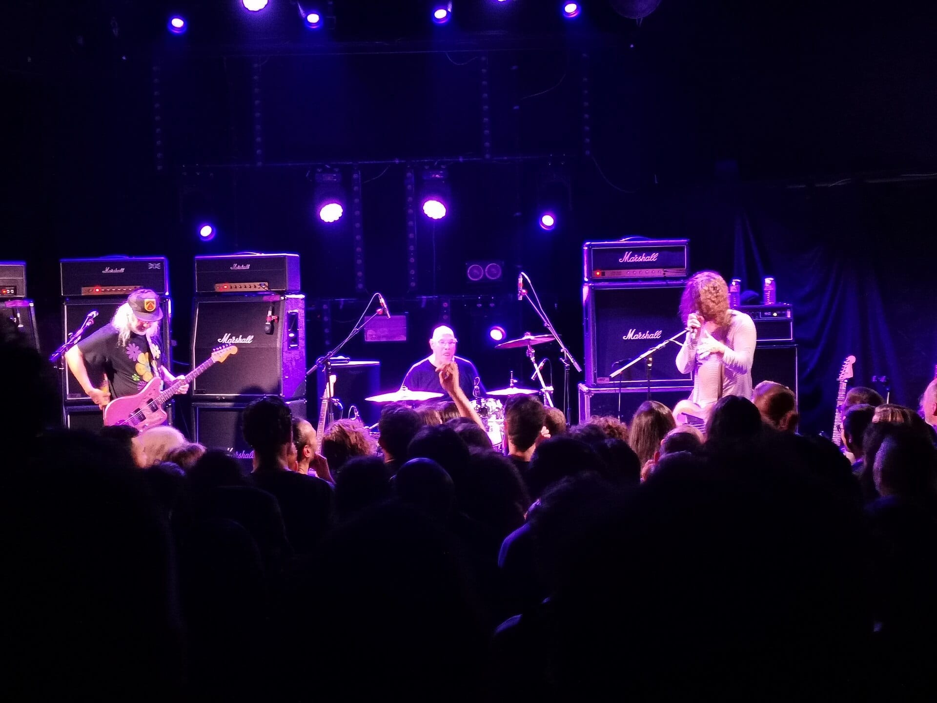 Dinosaur Jr. play a purple-lit club stage in front of stacked Marshall cabinets, J Mascis with white hair and a pink guitar at the left, the drummer centre and a singer at the right.