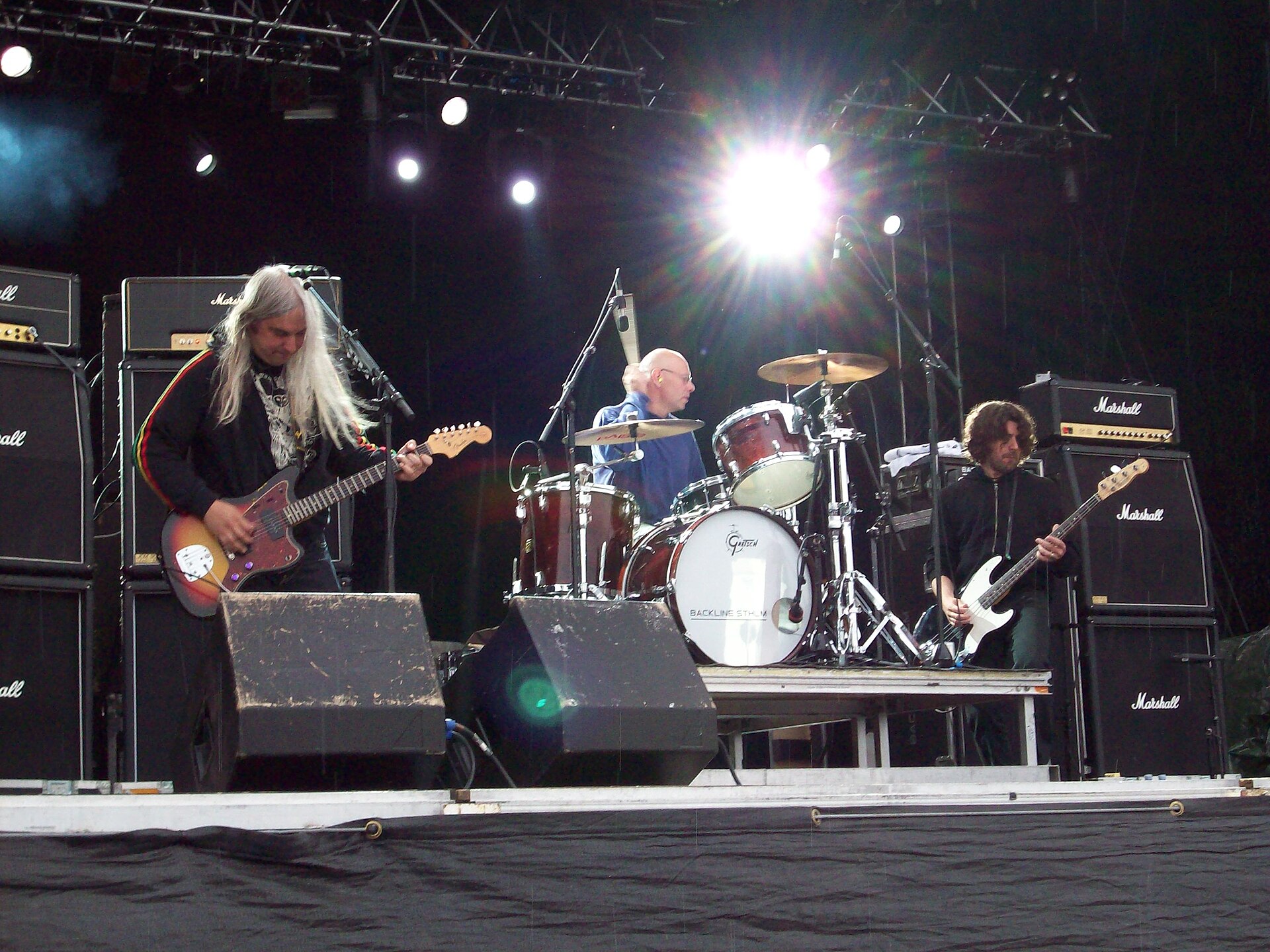 Dinosaur Jr. play an outdoor stage in front of walls of Marshall stacks, J Mascis with long white hair on the left, Murph at the kit and Lou Barlow on bass at the right.