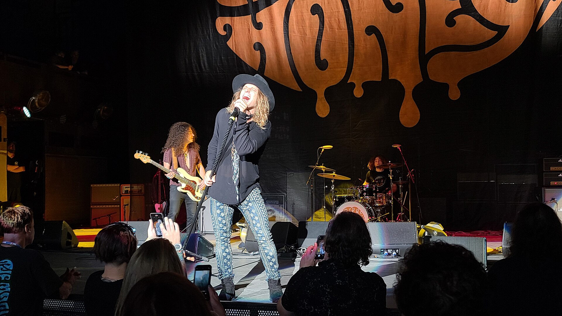 Marc LaBelle of Dirty Honey singing into a handheld microphone in a wide-brim hat and patterned jeans, the bassist and drummer behind him under a large dripping band banner