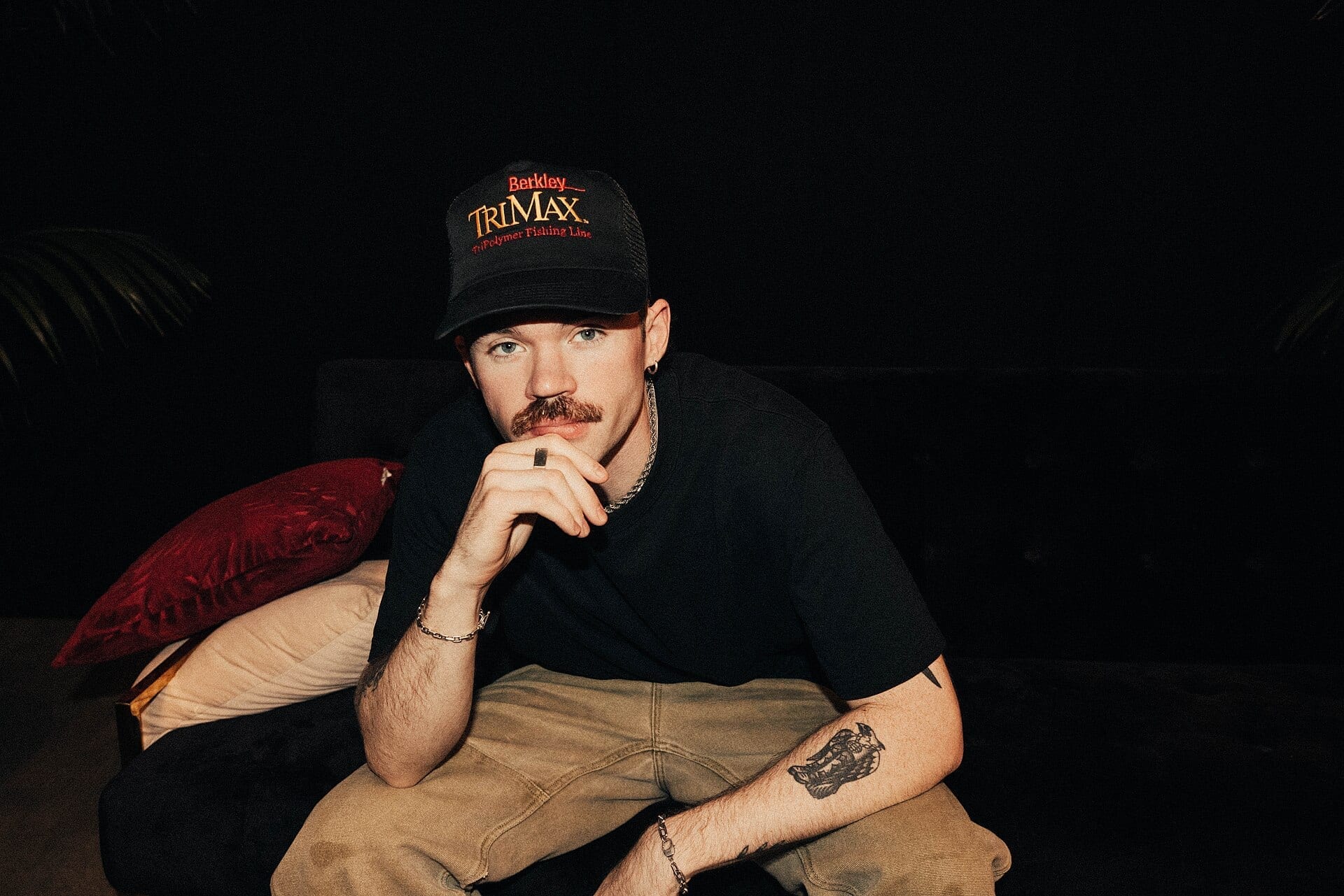 Dom Dolla sits for a portrait wearing a black trucker hat and black t-shirt, resting his chin on his hand.