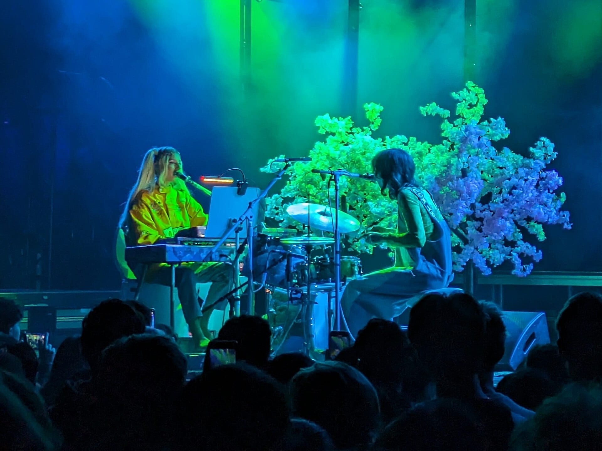 DOMi plays keyboard and sings while JD Beck plays drums, lit in green and blue with a floral backdrop.