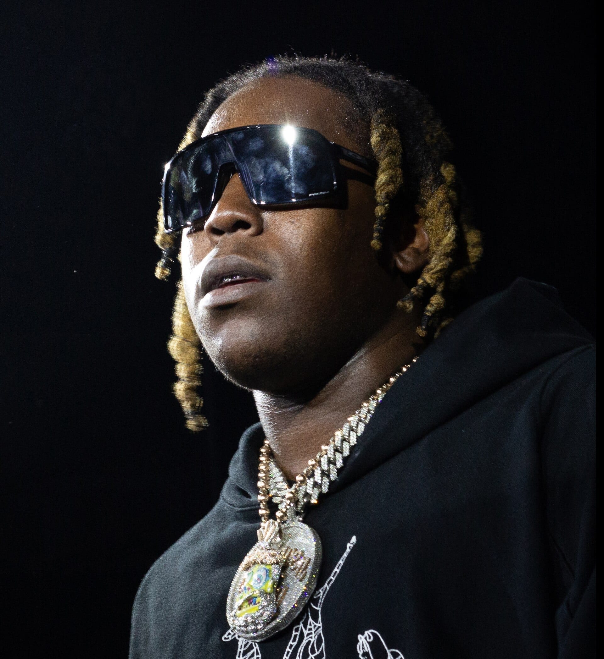 Don Toliver performs on stage in dramatic backlighting, wearing sunglasses and a diamond chain, with gold-tipped braids framing his face.