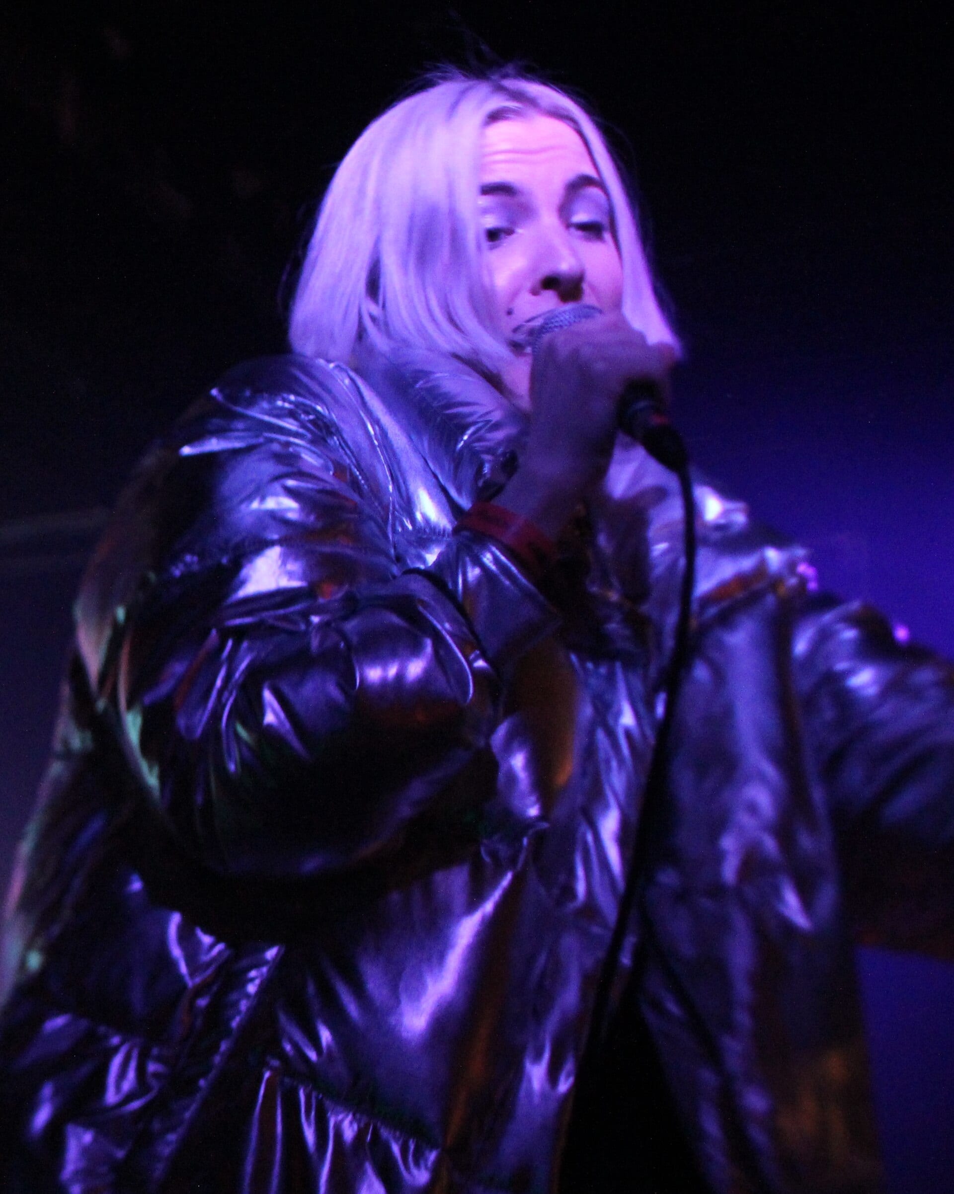 Dorian Electra sings into a handheld microphone under purple stage light, wearing a shiny silver puffer jacket with a red neck strap and platinum blonde hair falling to the shoulders.