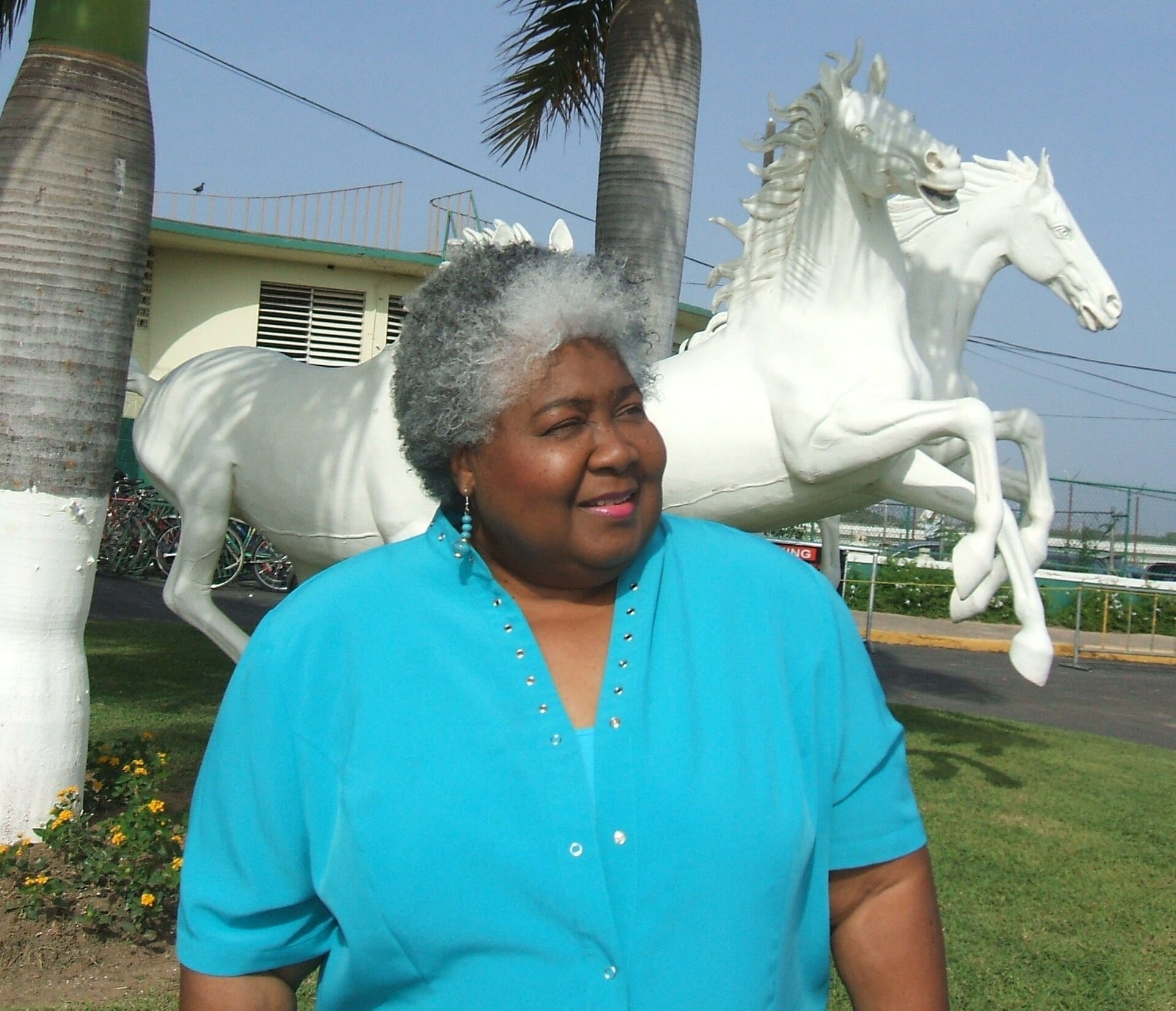 Dorothy Moore smiles in a bright turquoise blouse, standing in front of two white statues of galloping horses and a row of palm trees.