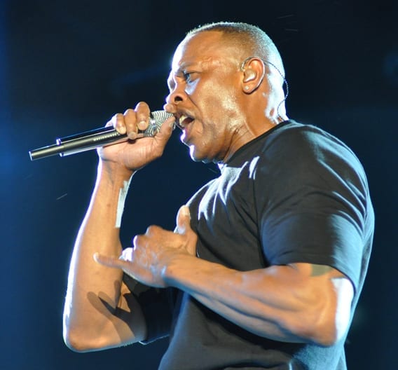 Dr. Dre sings into a handheld microphone under blue stage light, wearing a black T-shirt and an in-ear monitor.