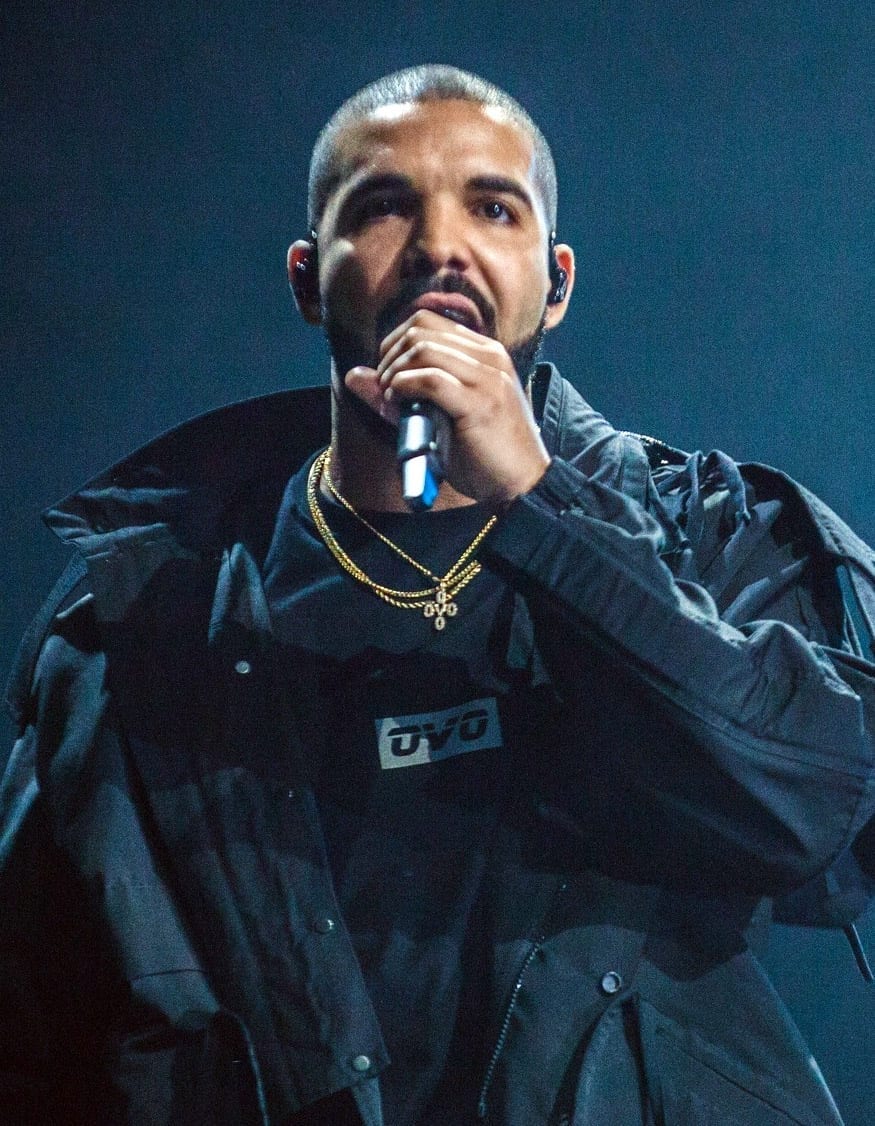 Drake sings into a handheld microphone on a dim stage, wearing a dark jacket over an OVO T-shirt and layered gold chains.