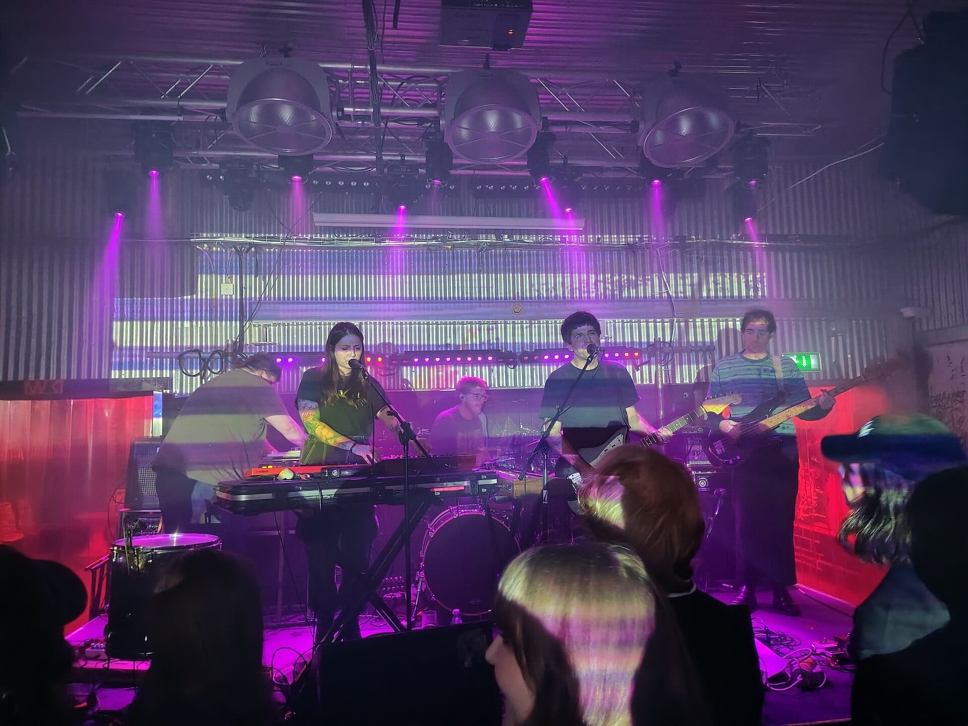 The band Dummy performs on a purple-lit stage, with members on keyboards, guitar and bass under a corrugated metal backdrop.