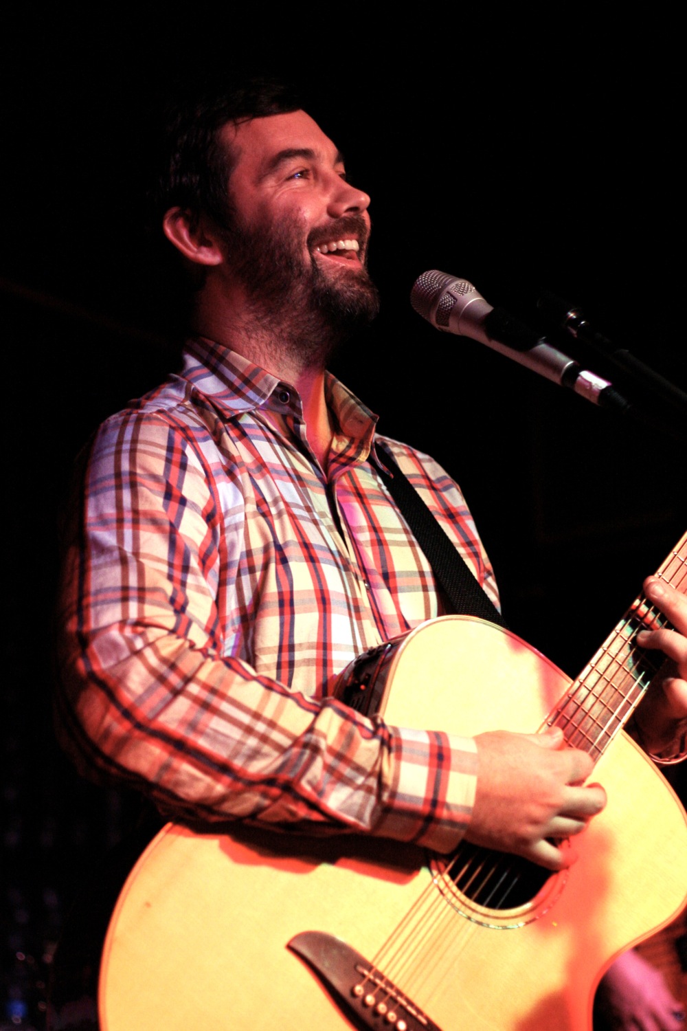 Duncan Sheik laughs mid-song at a club microphone, bearded and in a plaid shirt, holding a pale acoustic guitar.