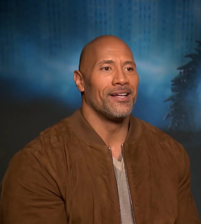 Dwayne Johnson, wearing a brown jacket, speaks during an interview against a blurred blue backdrop.
