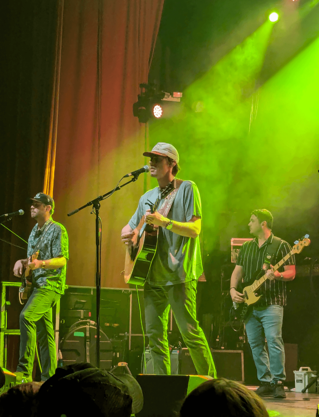 Dylan Gossett sings into a microphone while playing an acoustic guitar, wearing a ball cap, with his band performing behind him under green stage lighting.