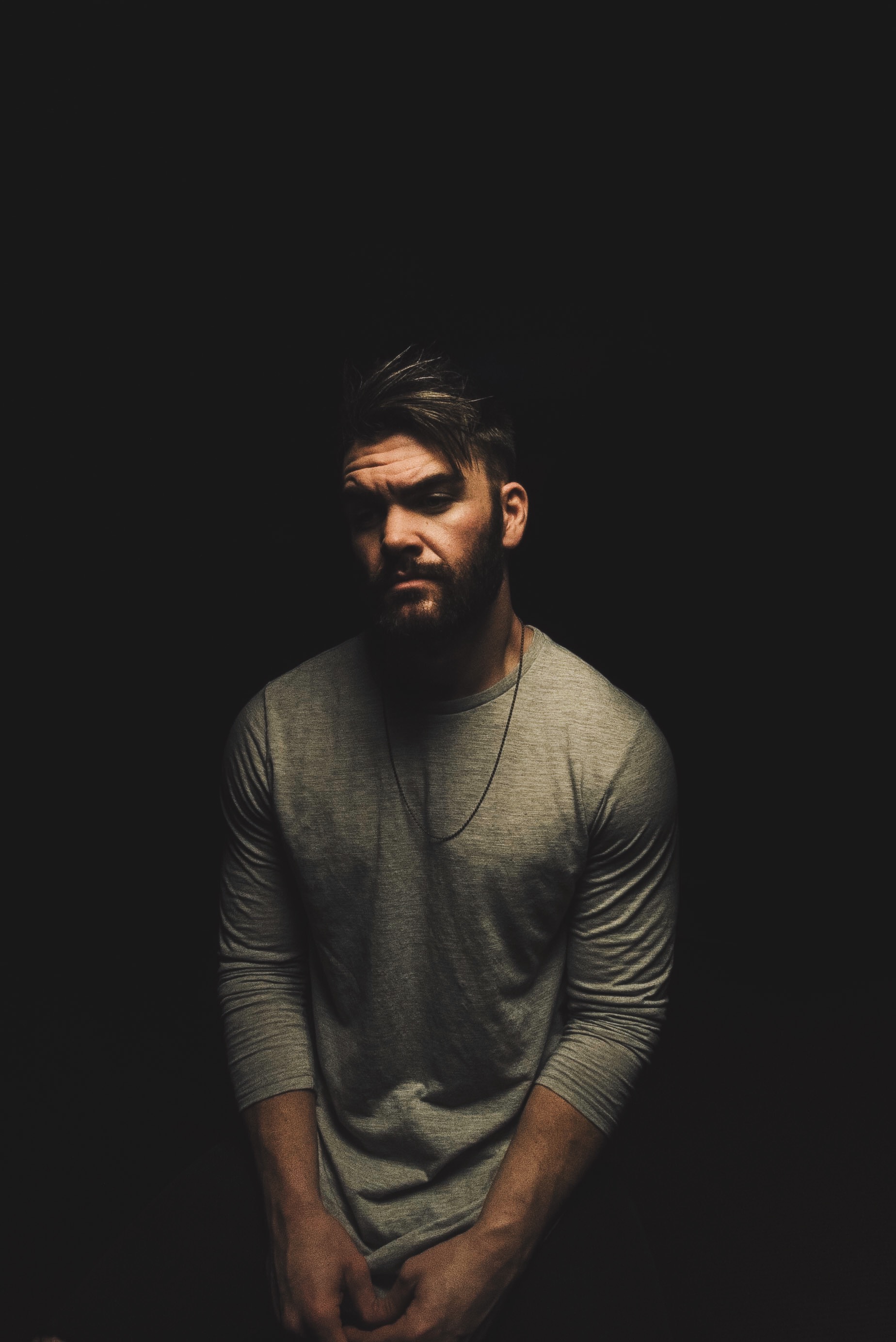 Dylan Scott stands against a black background in a low-lit studio portrait, in a plain grey long-sleeved shirt and a thin chain, looking straight at the camera.