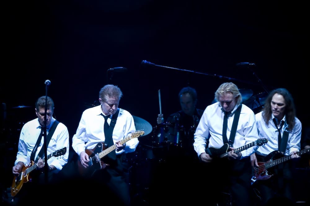 The Eagles perform on a dark stage in matching white shirts and black ties, playing electric guitars with a drummer visible behind them.