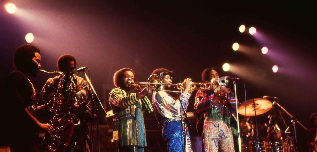 Members of Earth, Wind & Fire's horn section play trumpets, trombone, and saxophone side by side on a brightly lit stage, wearing colorful patterned outfits.
