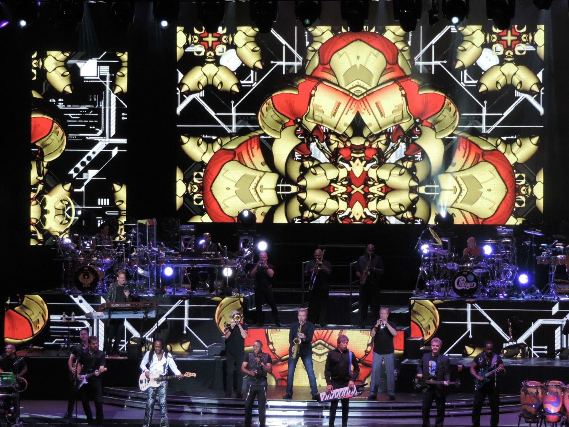 Members of Earth, Wind & Fire and Chicago perform together on a large stage lit with kaleidoscopic red and gold graphics on the screens behind them.