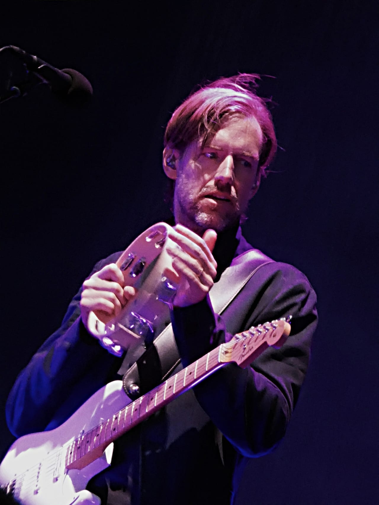 Ed O'Brien tunes a white electric guitar on a dimly lit stage, bathed in purple and blue light.
