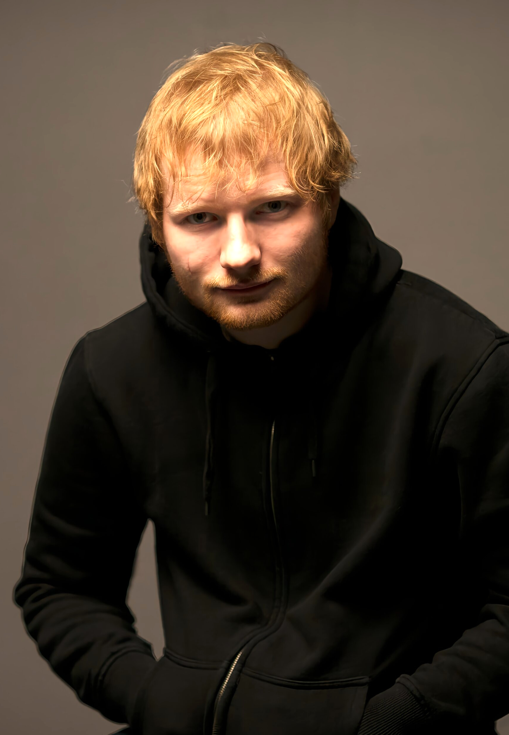 Ed Sheeran looks straight at the camera in a dark shirt, close cropped against a plain background.