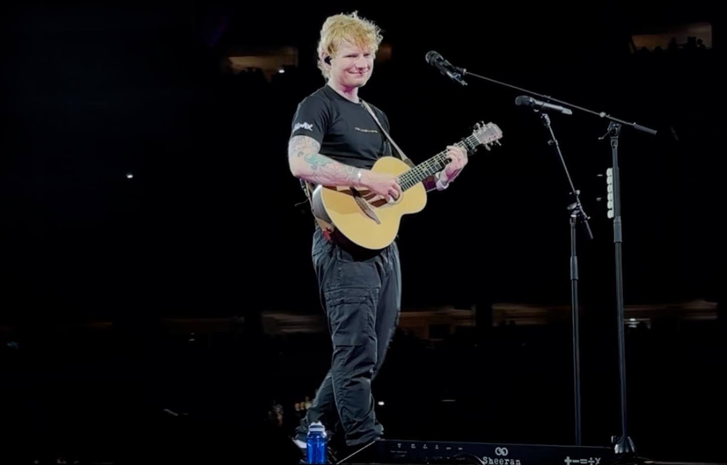 Ed Sheeran stands alone on a dark stadium stage with an acoustic guitar, in a black T-shirt and dark cargo trousers, two mic stands beside him.
