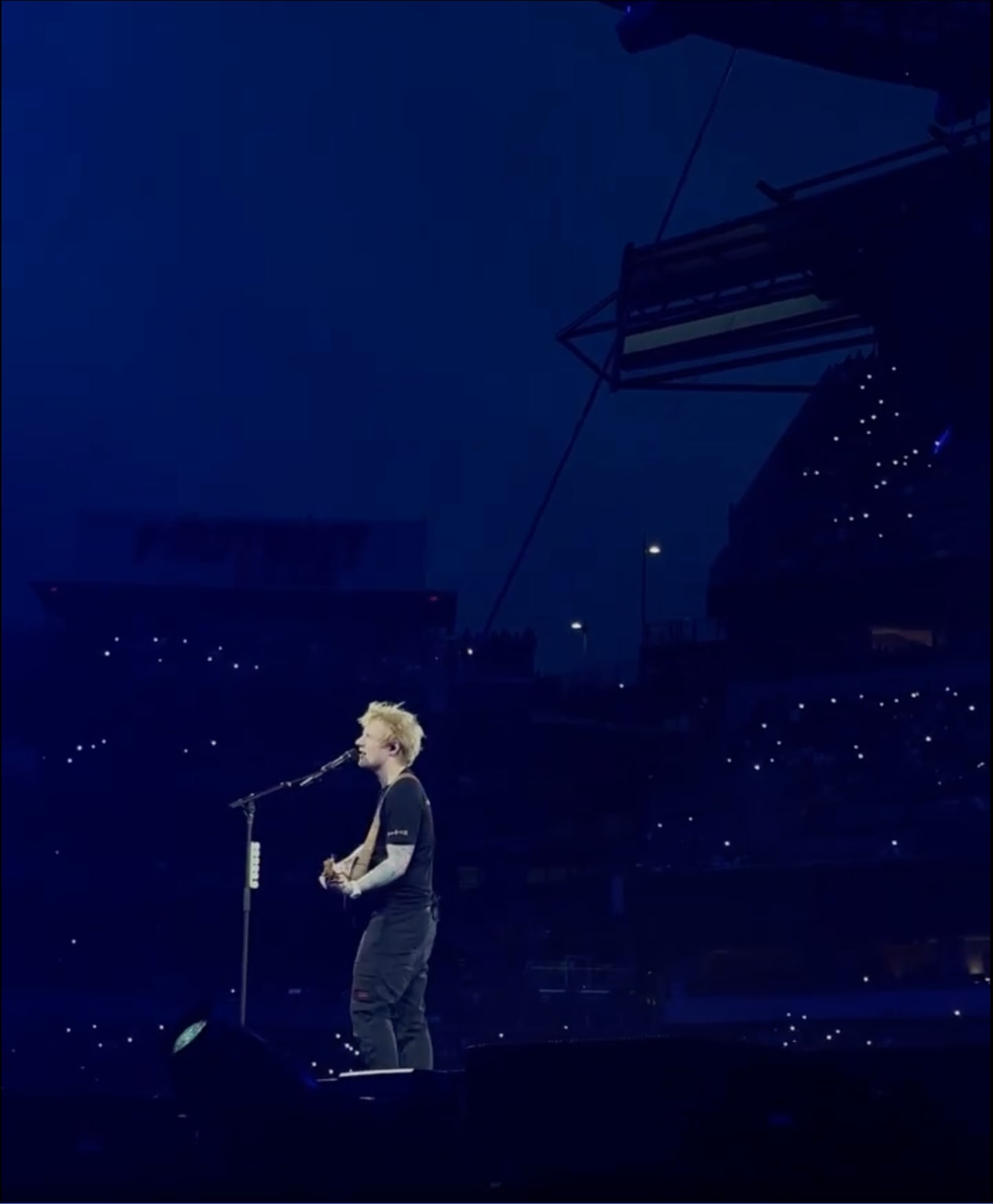 A red-haired singer stands alone with a guitar at a microphone stand on a stadium stage at dusk, phone lights scattered through the dark stands behind him