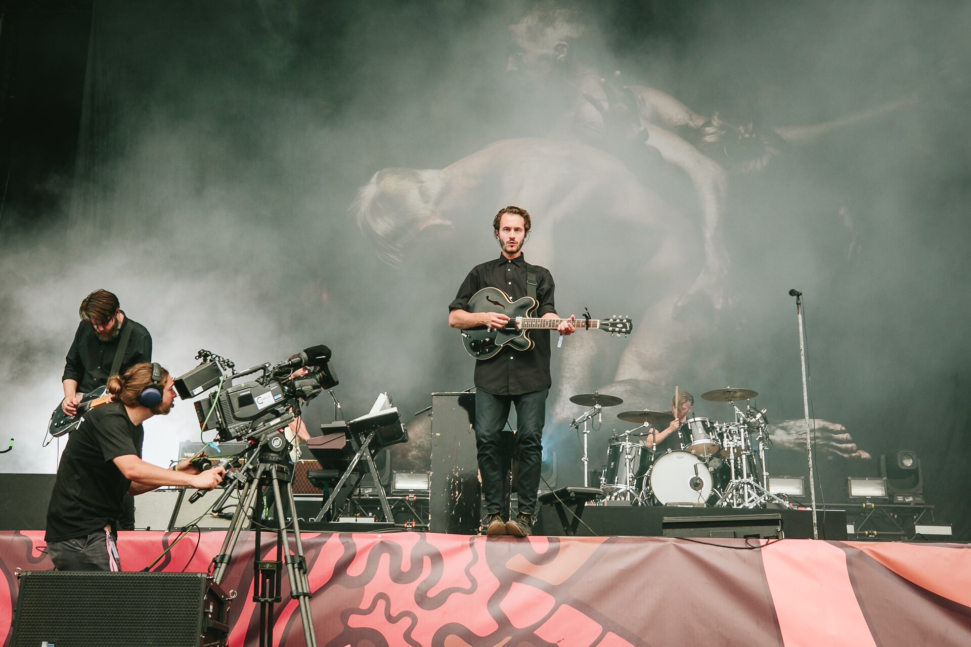 Editors on a smoke-filled festival stage, the singer centre in a black shirt with a hollow-body guitar, a guitarist to the left and drummer to the right, a huge animal image on the backdrop behind them.