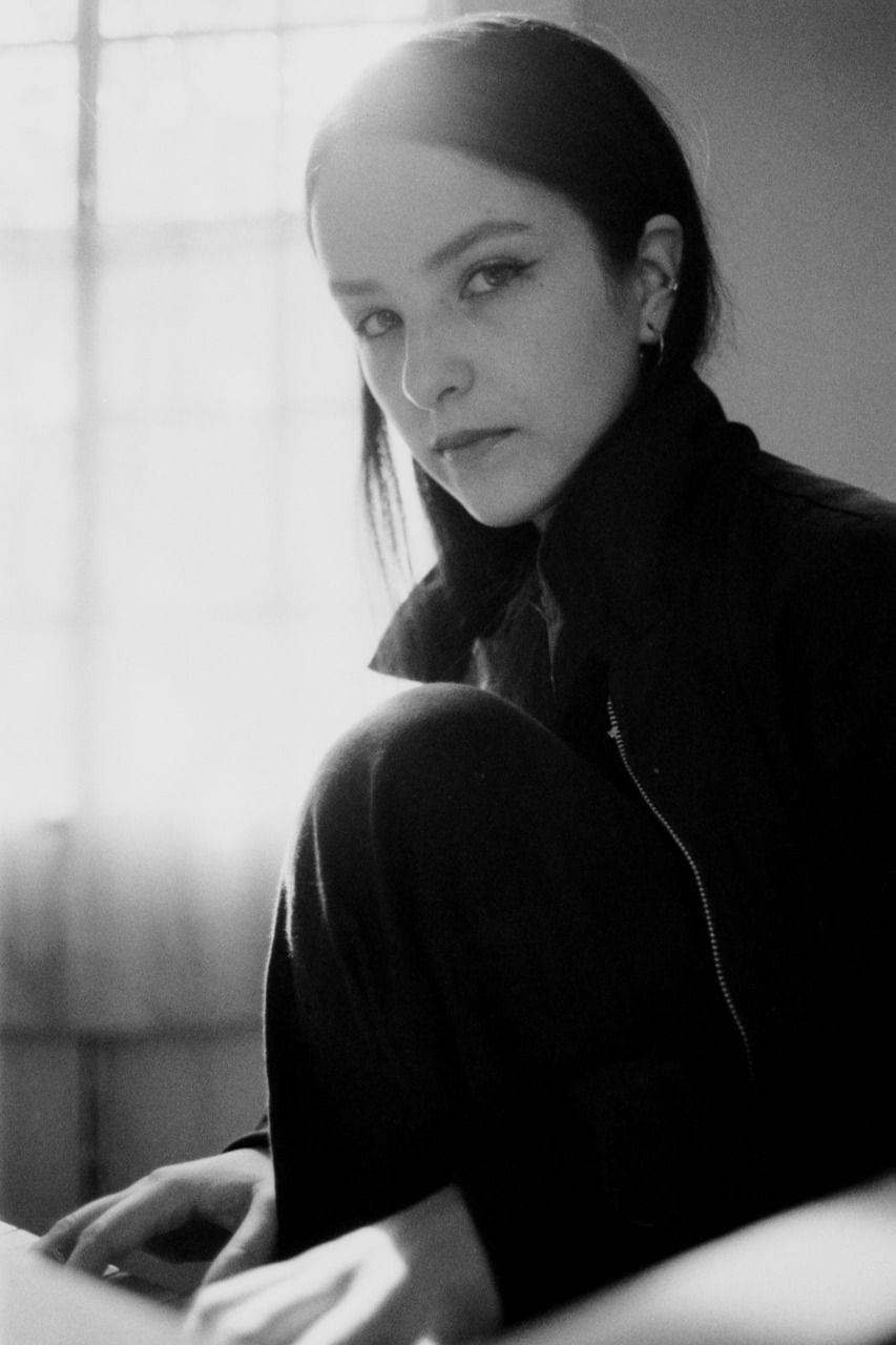 A black-and-white portrait of Ela Minus sitting by a window in a black zip-up jacket, hugging her knee and looking toward the camera.