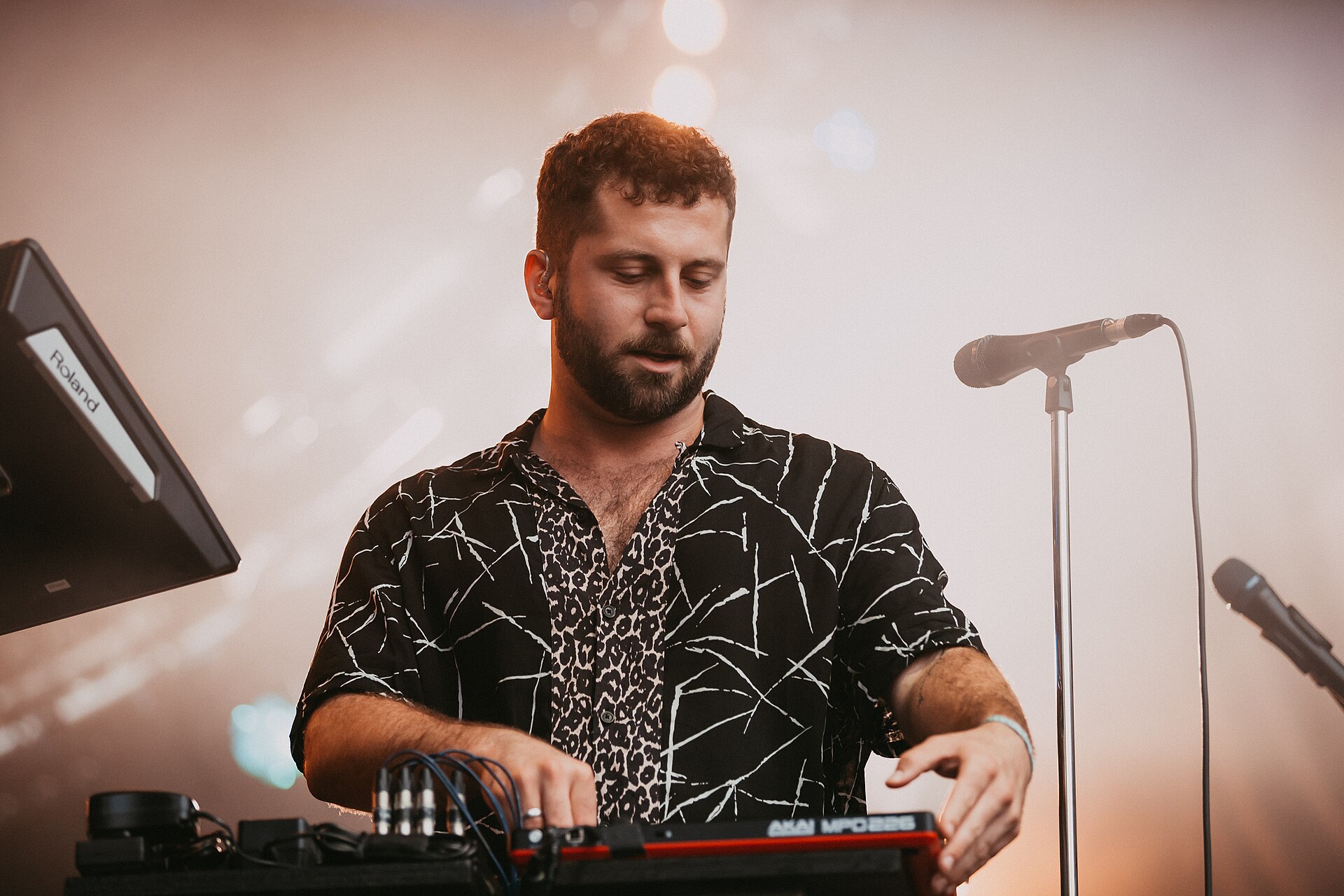 Elderbrook works over a bank of synths and drum machines on a festival stage with a microphone stand beside him.