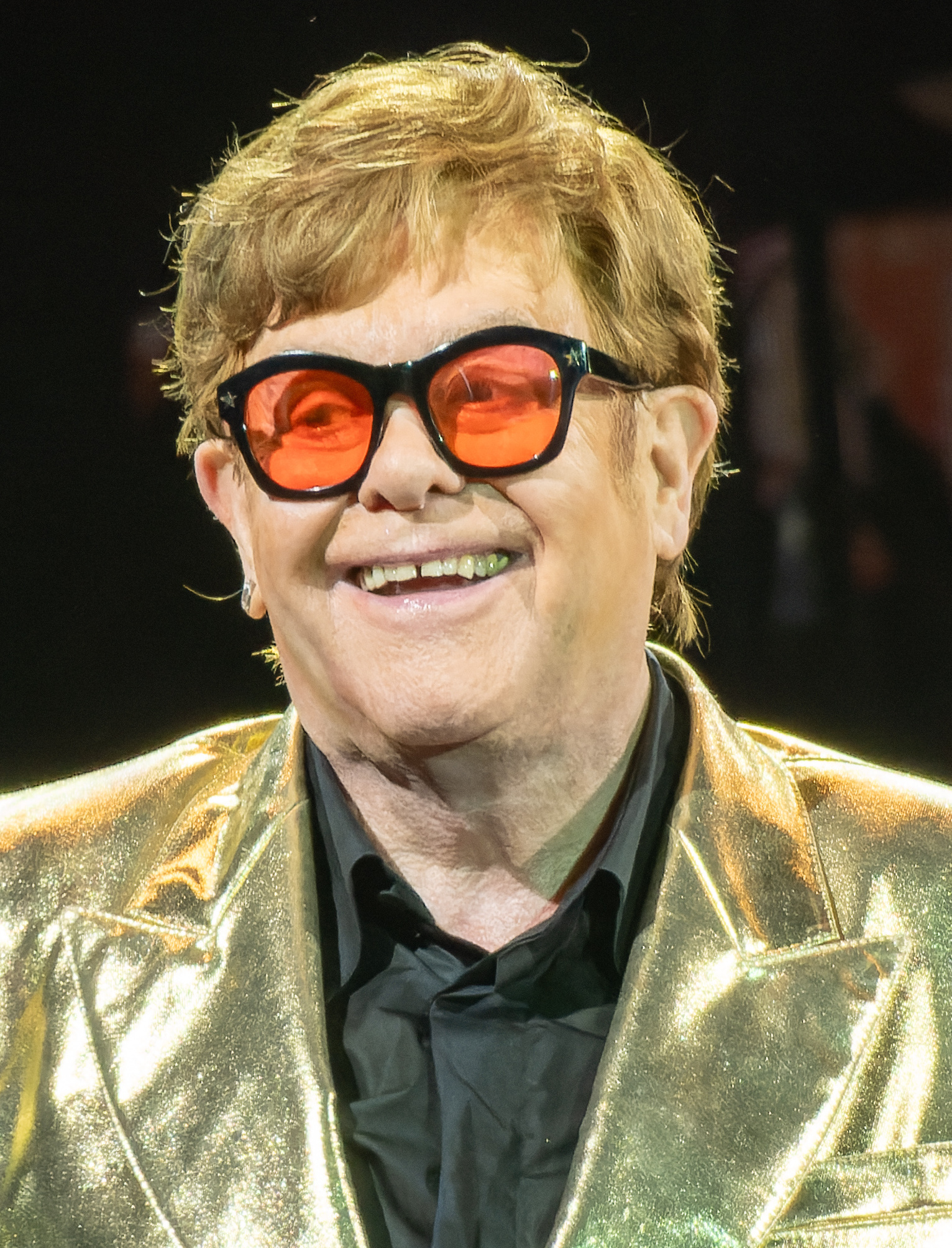 Elton John smiles in close-up wearing orange-lensed glasses with black frames and a gold lame jacket over a black shirt.