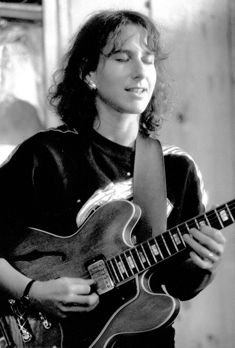 A black and white photograph of Emily Remler playing a hollow-body electric guitar with her eyes closed, in a dark sweatshirt with a wide strap over one shoulder.
