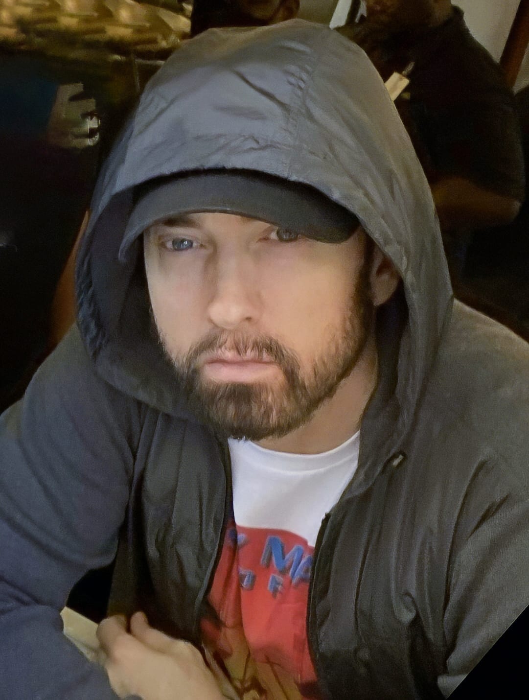 Eminem looks toward the camera wearing a gray hooded jacket and a baseball cap, with a full beard.