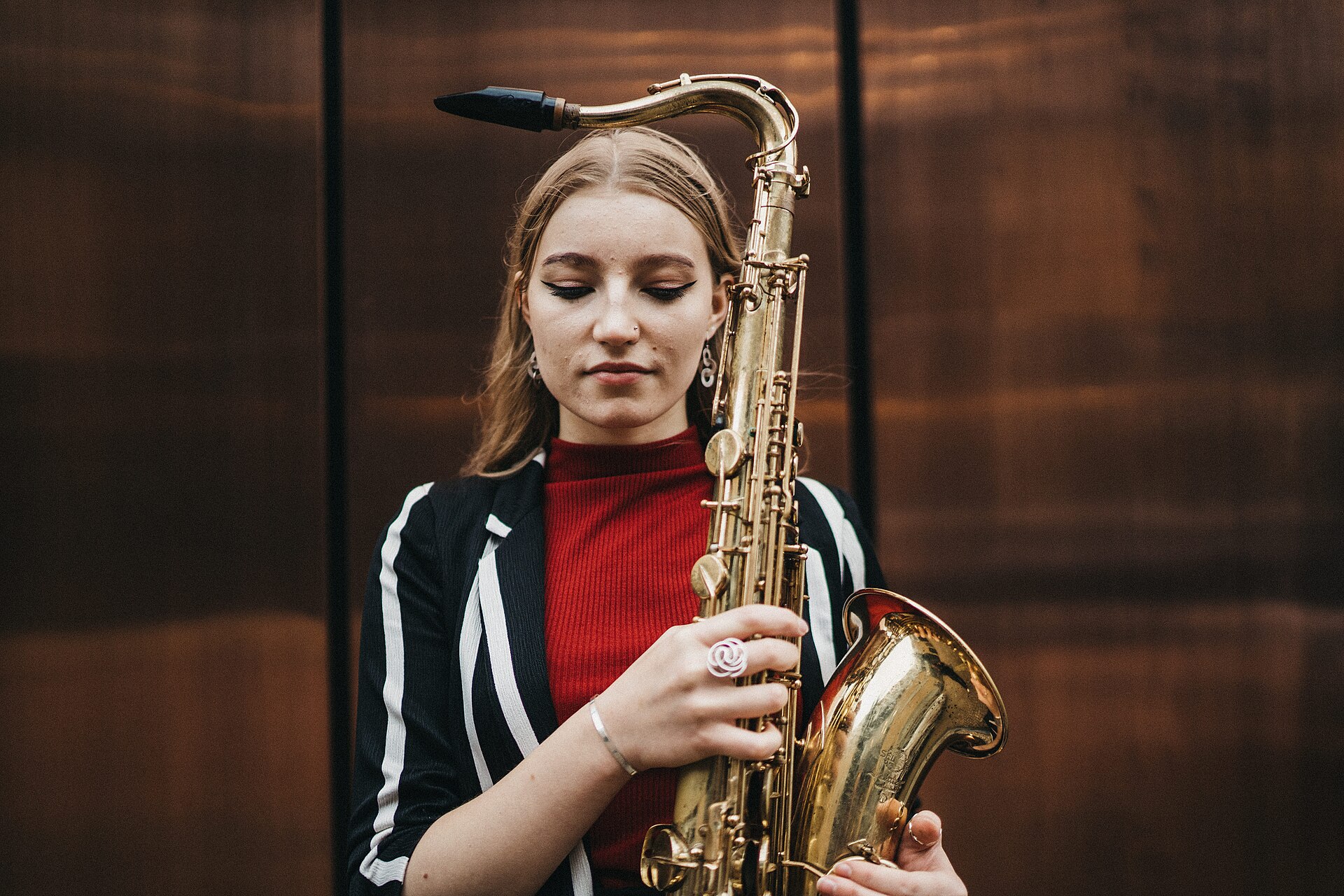 Emma Rawicz holding a tenor saxophone upright in front of her, looking down, in a striped jacket over a red top against a dark copper wall