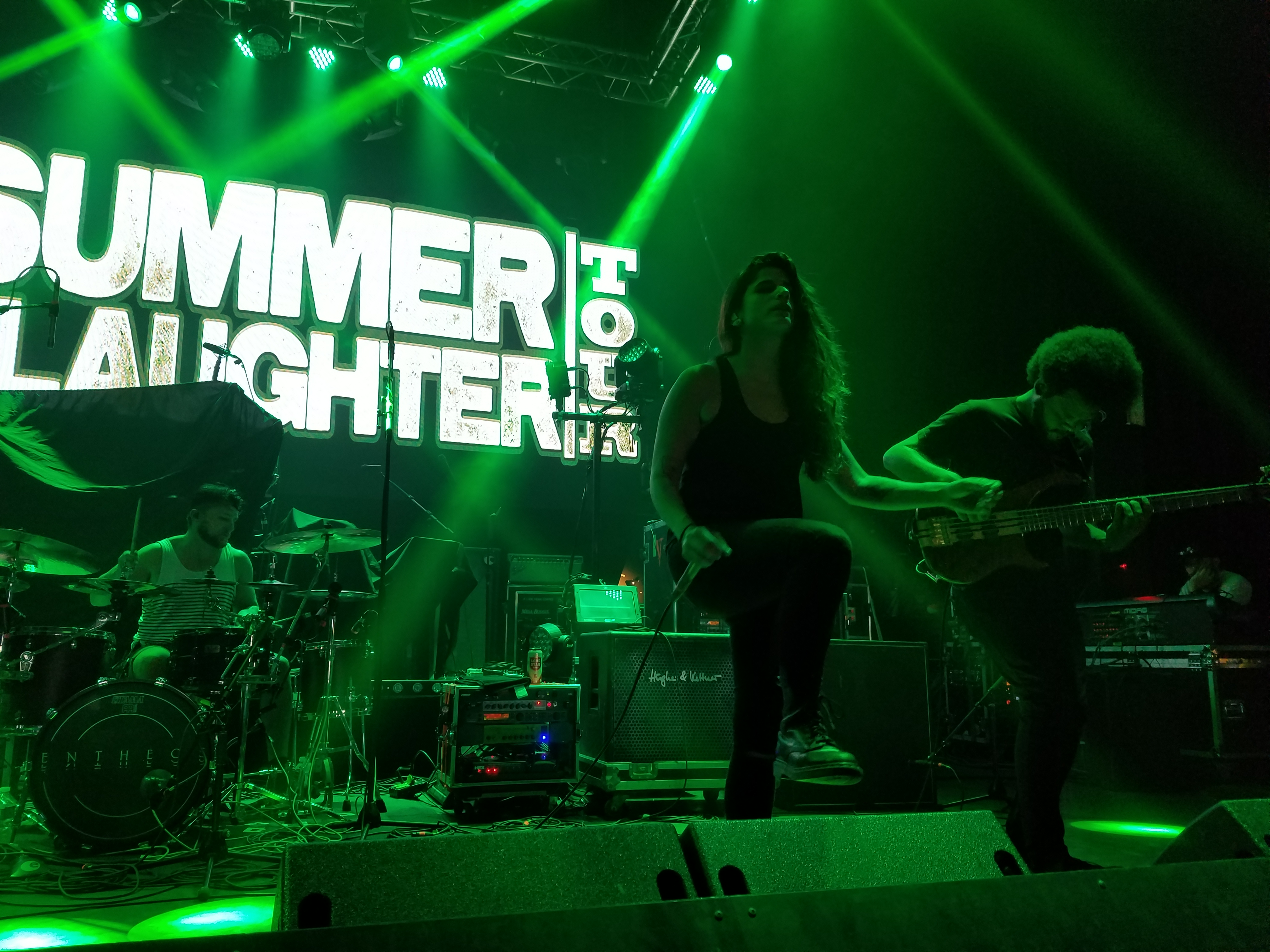Entheos play under green stage lights in front of a Summer Slaughter Tour banner, the singer centre stage in a black tank top with a bassist beside her and a drummer behind.