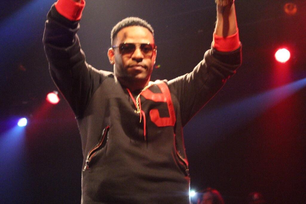Eric Bellinger raises both arms above his head on stage in sunglasses and a black and red sweatshirt.