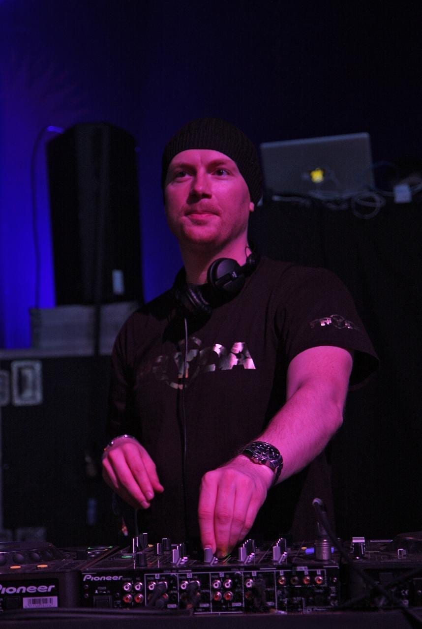 Eric Prydz stands behind a DJ mixer in a beanie with headphones around his neck, lit in pink and purple.