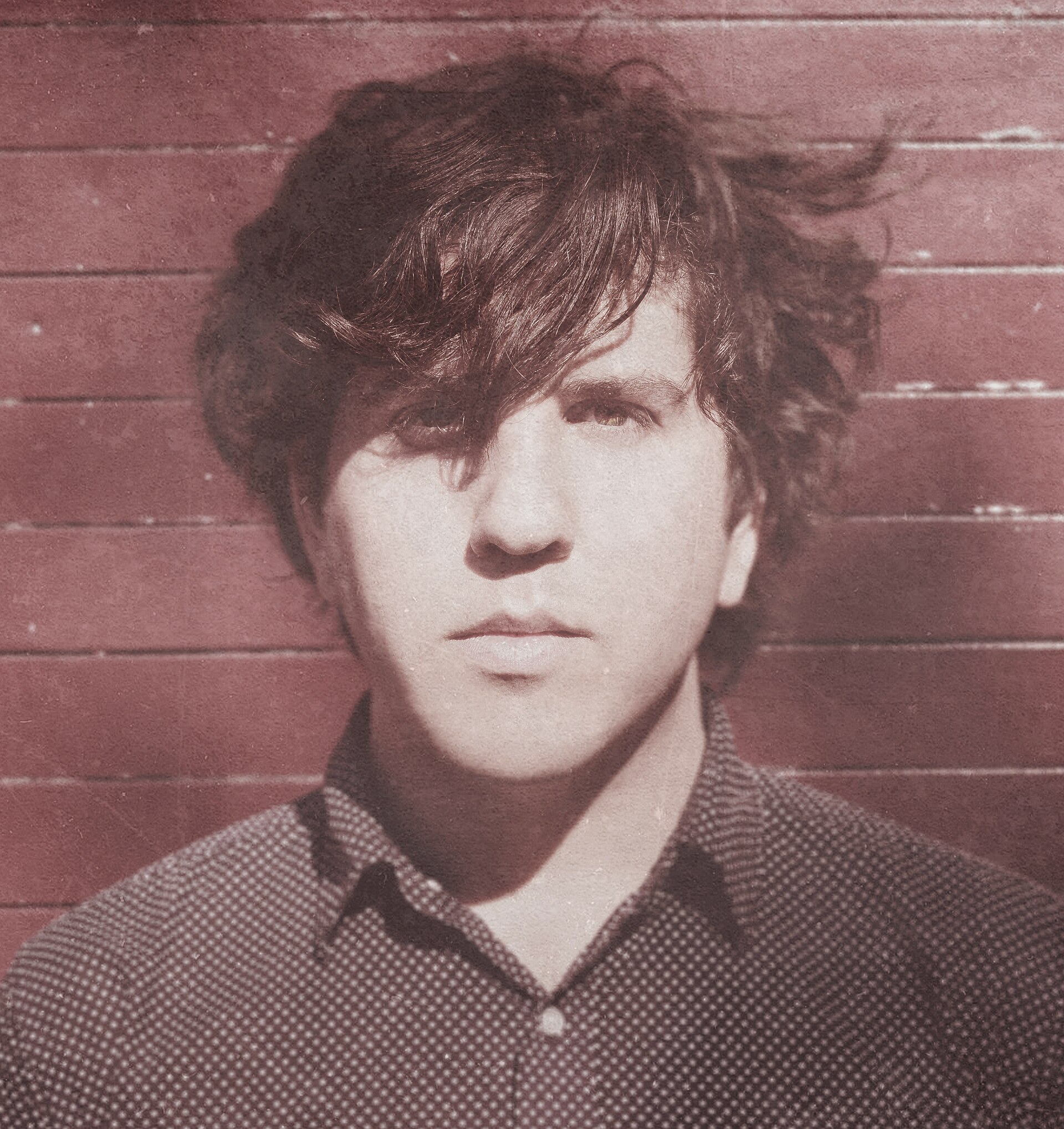 A head-and-shoulders portrait of Eric Slick in a dark patterned button-up shirt, hair falling across one eye, against a red painted wall.
