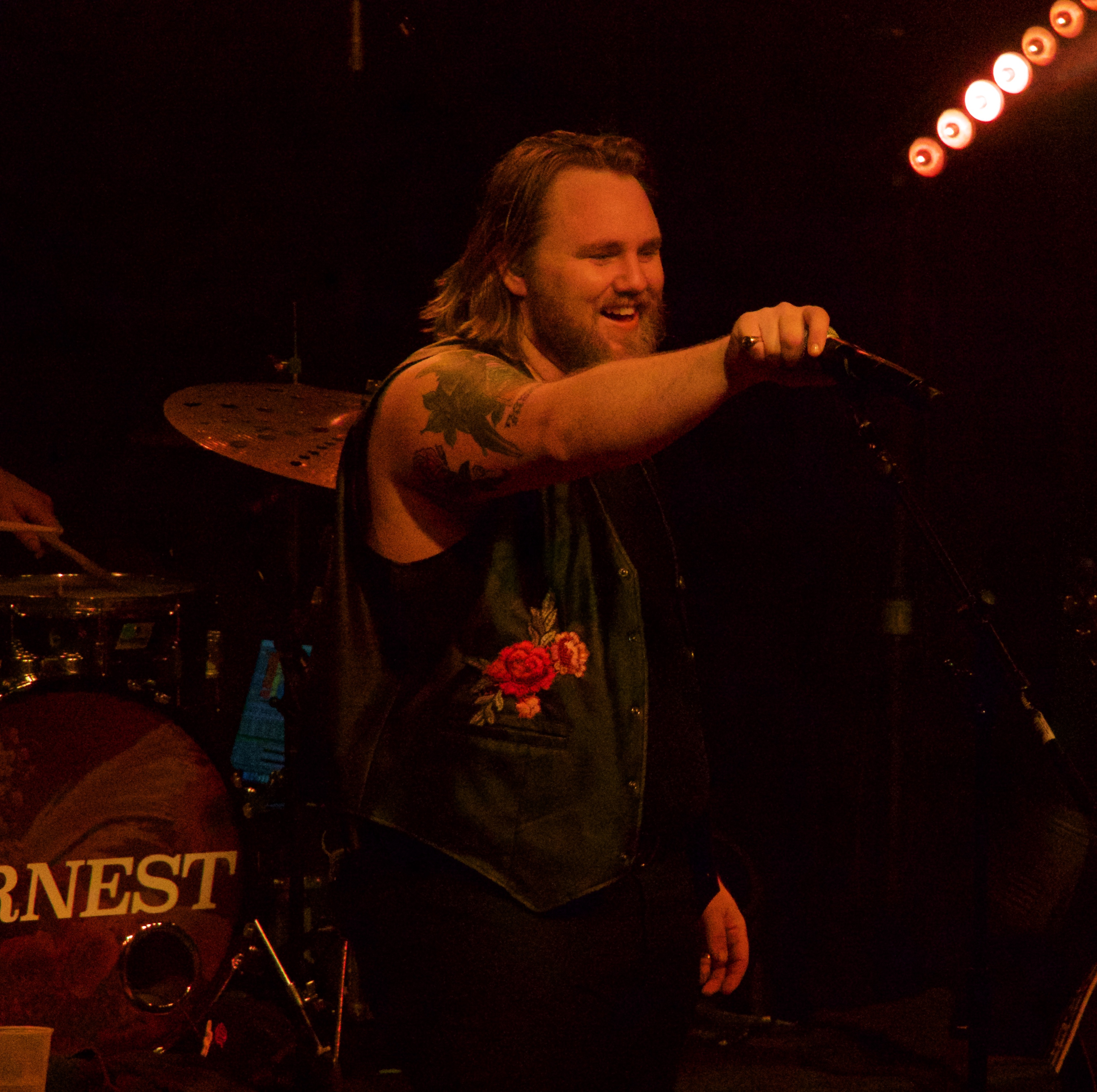 ERNEST grins and points his microphone out at the crowd in a black waistcoat embroidered with roses, a bass drum head reading ERNEST behind him.