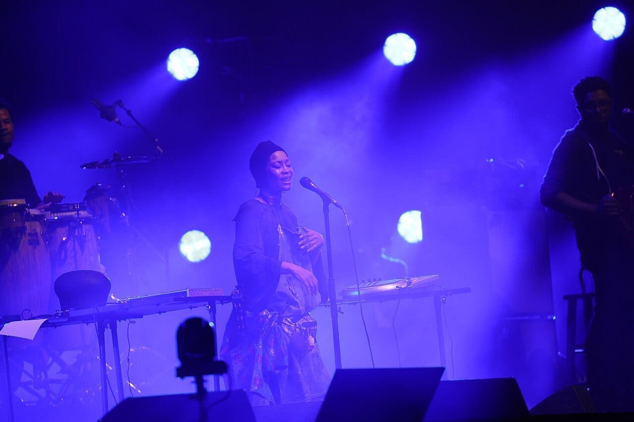 Erykah Badu smiles at a microphone stand behind a keyboard rig in a tall black cap, washed in blue light and haze, a percussionist and a guitarist either side of her.