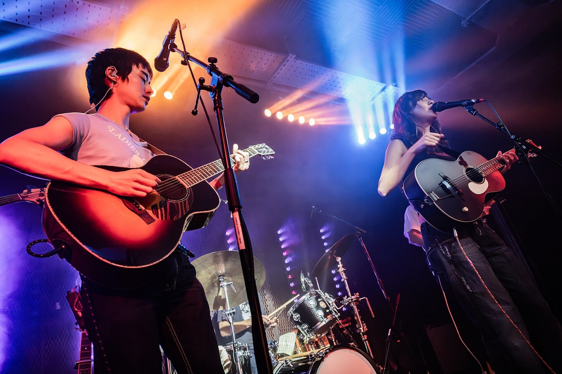 The two members of Esme Emerson perform on stage under green and pink stage lights, one playing an acoustic guitar and singing at a microphone, the other singing while playing a smaller guitar.