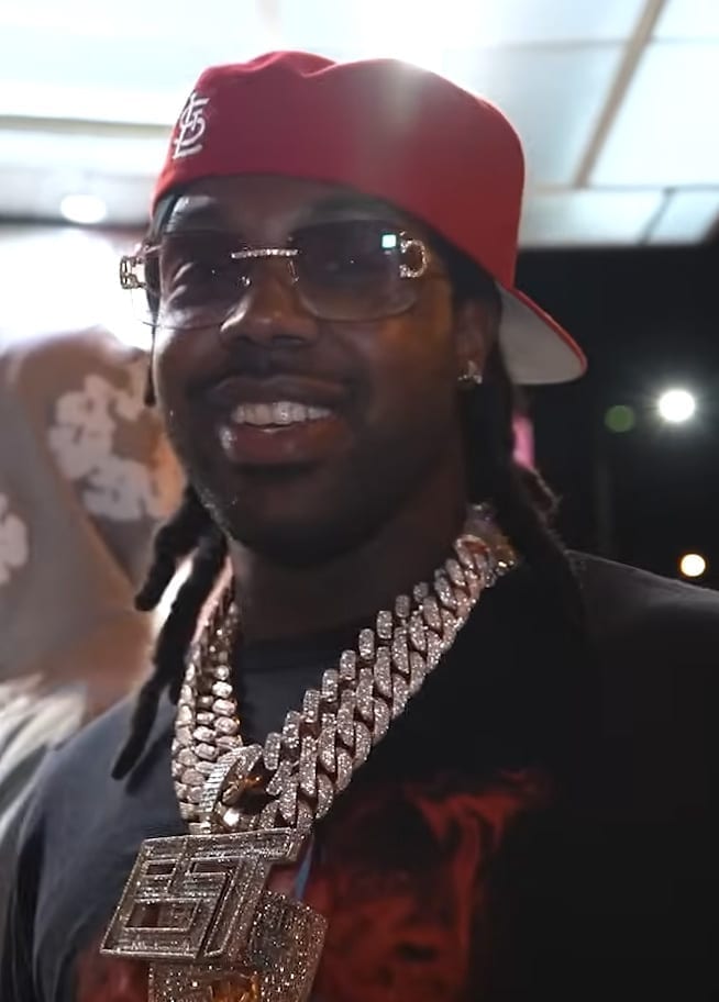 EST Gee, wearing a red cap, tinted glasses and a large diamond chain, smiles during an interview.