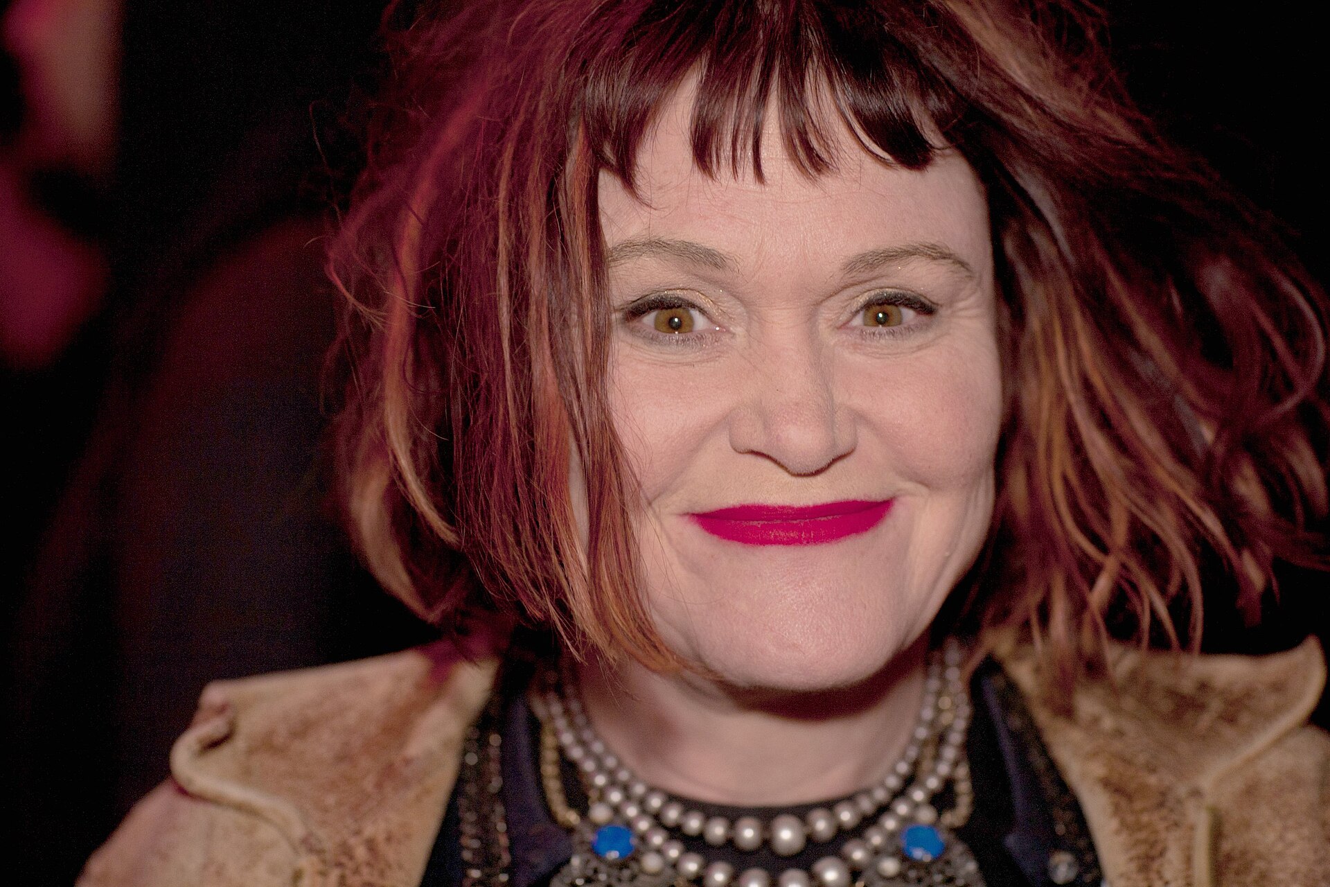 Exene Cervenka smiles at the camera in close up, dark red hair falling around her face over a shearling collar.