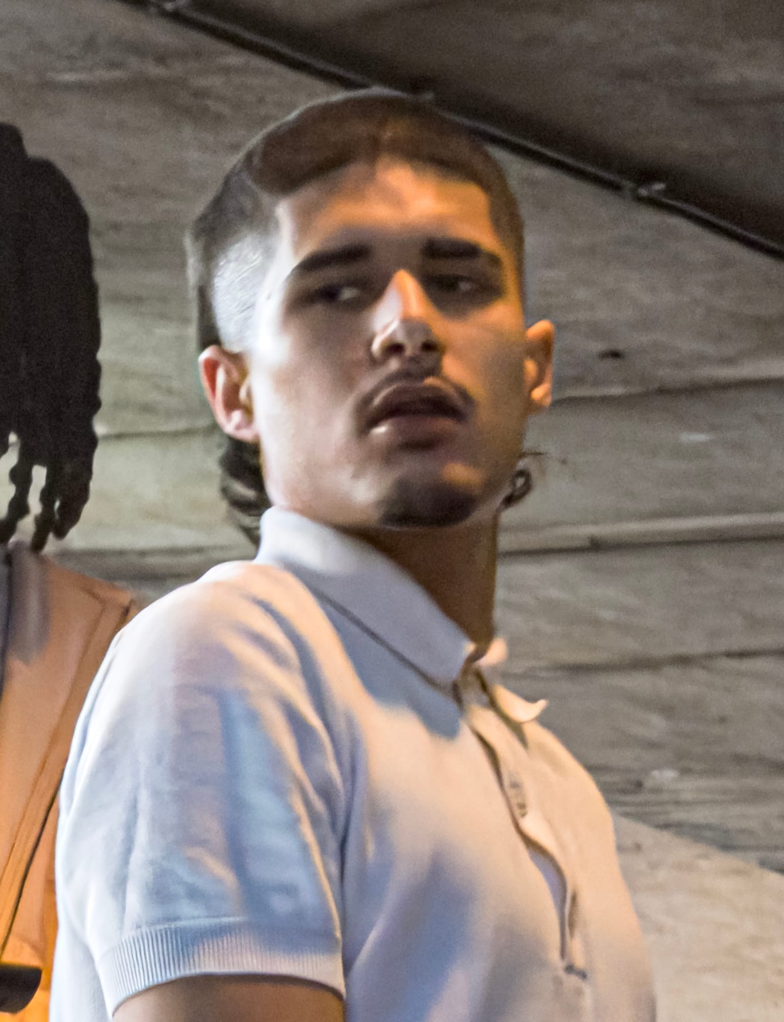 Fakemink, wearing a light-colored polo shirt, looks over his shoulder in a dim, low-ceilinged space, his hair in braids.