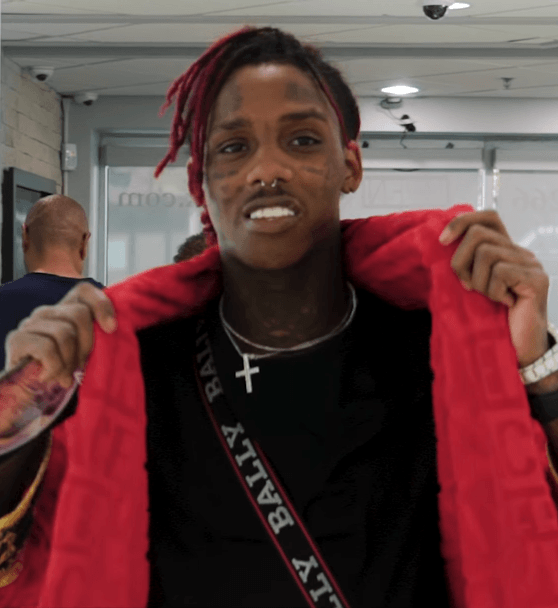 Famous Dex holds open the collar of a red hoodie and looks into the camera, red-dyed dreadlocks tied up and small tattoos across his cheeks.