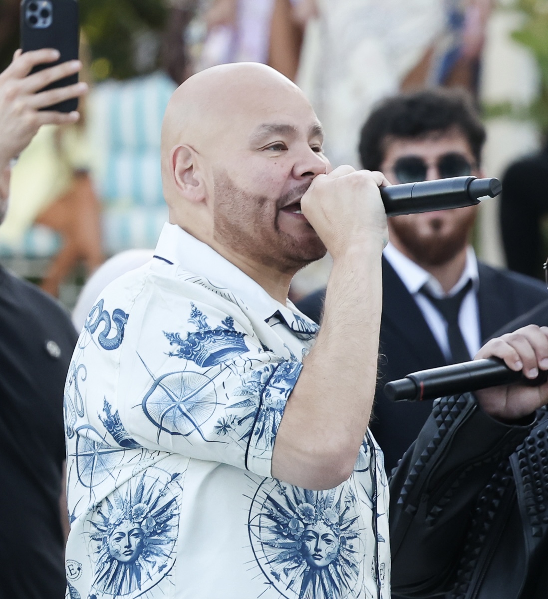 Fat Joe speaks into a handheld microphone outdoors in a white shirt printed with blue suns and crowns, a phone camera raised behind him.