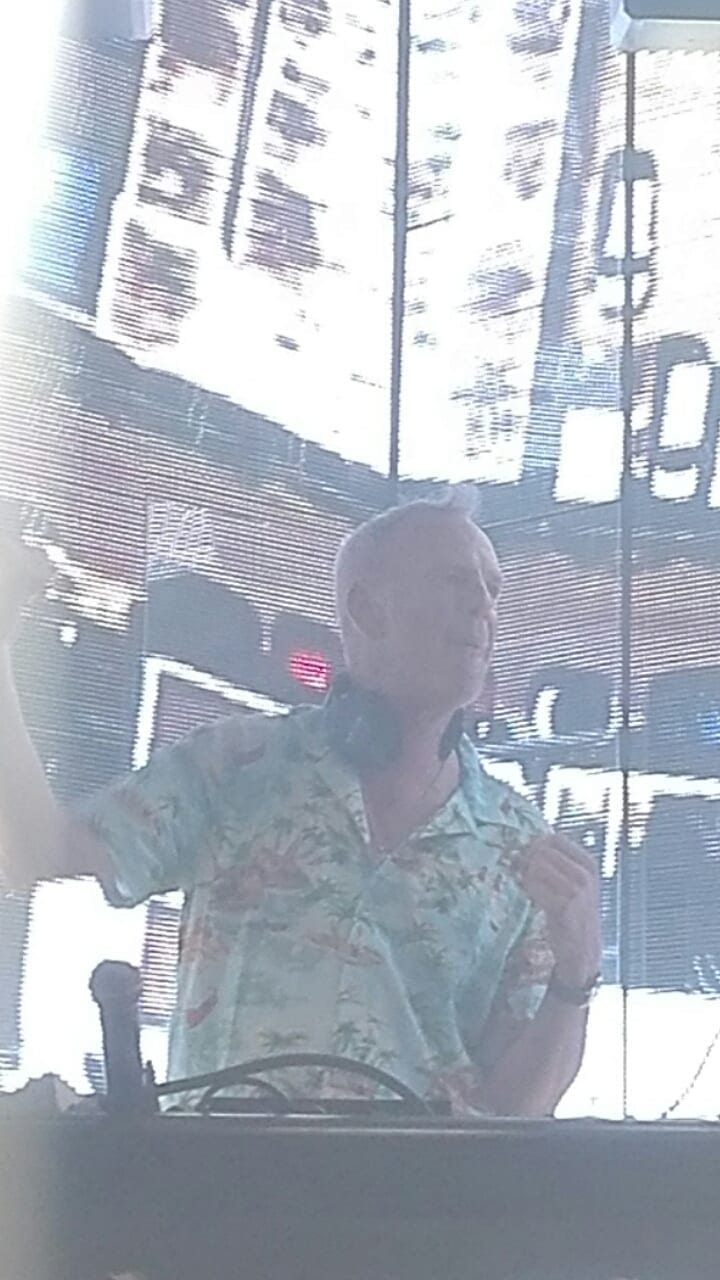 Fatboy Slim, in a pale green Hawaiian shirt and headphones around his neck, DJs in front of a bright video screen backdrop.