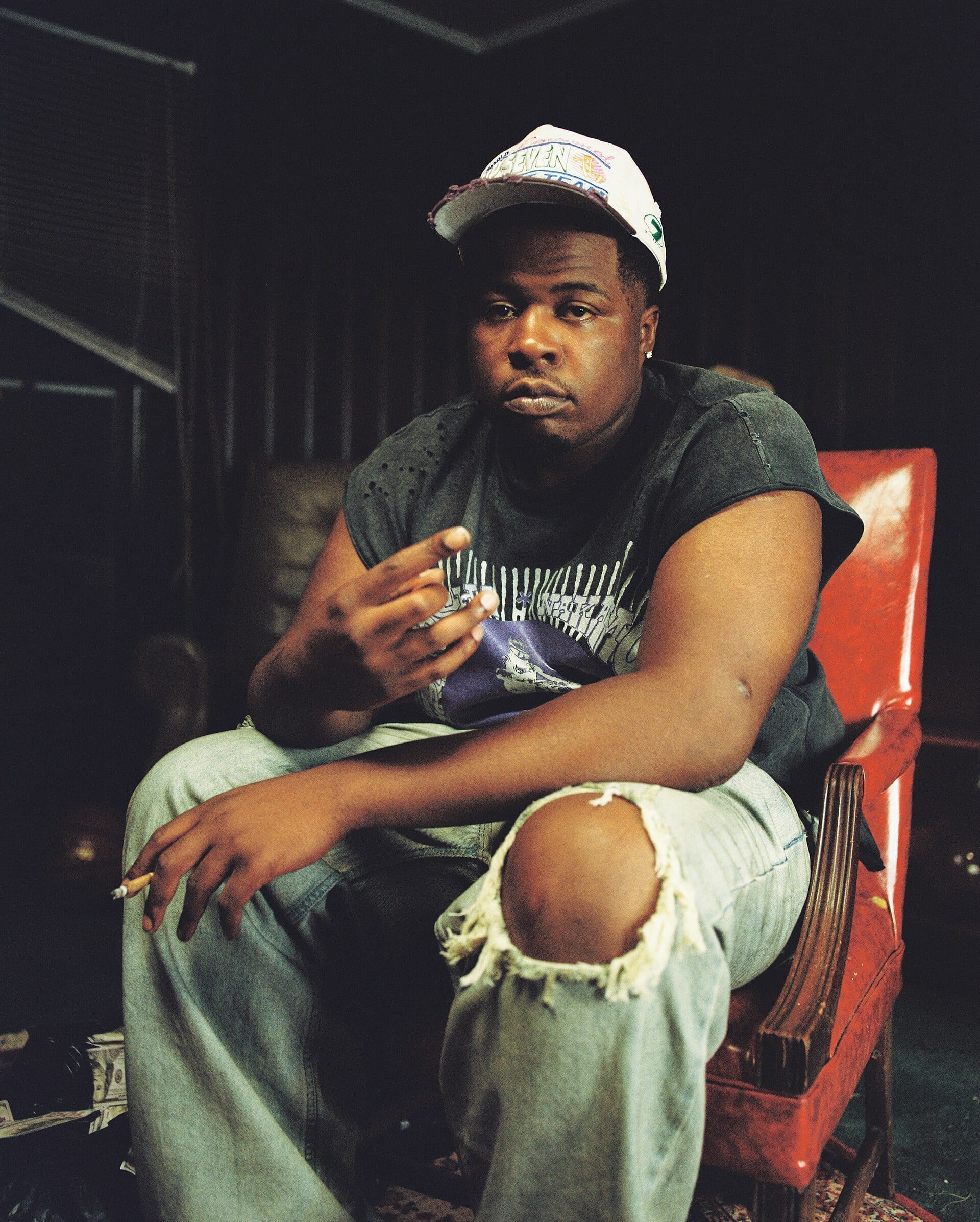 Fatt Smaxk sits in a red armchair wearing a white cap and jeans ripped at the knee, holding a lit cigarette and looking at the camera.