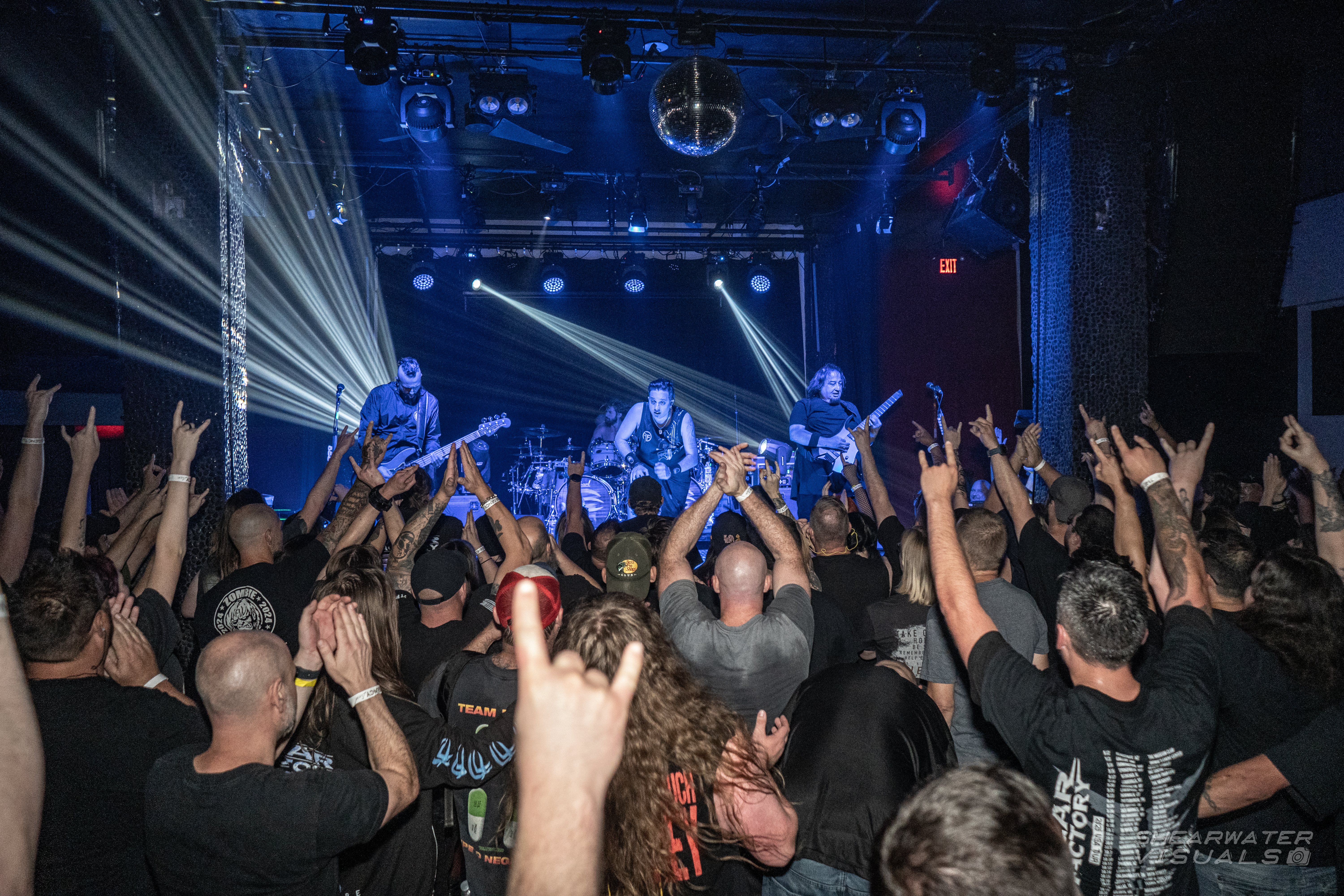 Fear Factory play a club stage in white beams of light, seen from the back of a packed floor of fans throwing horns.