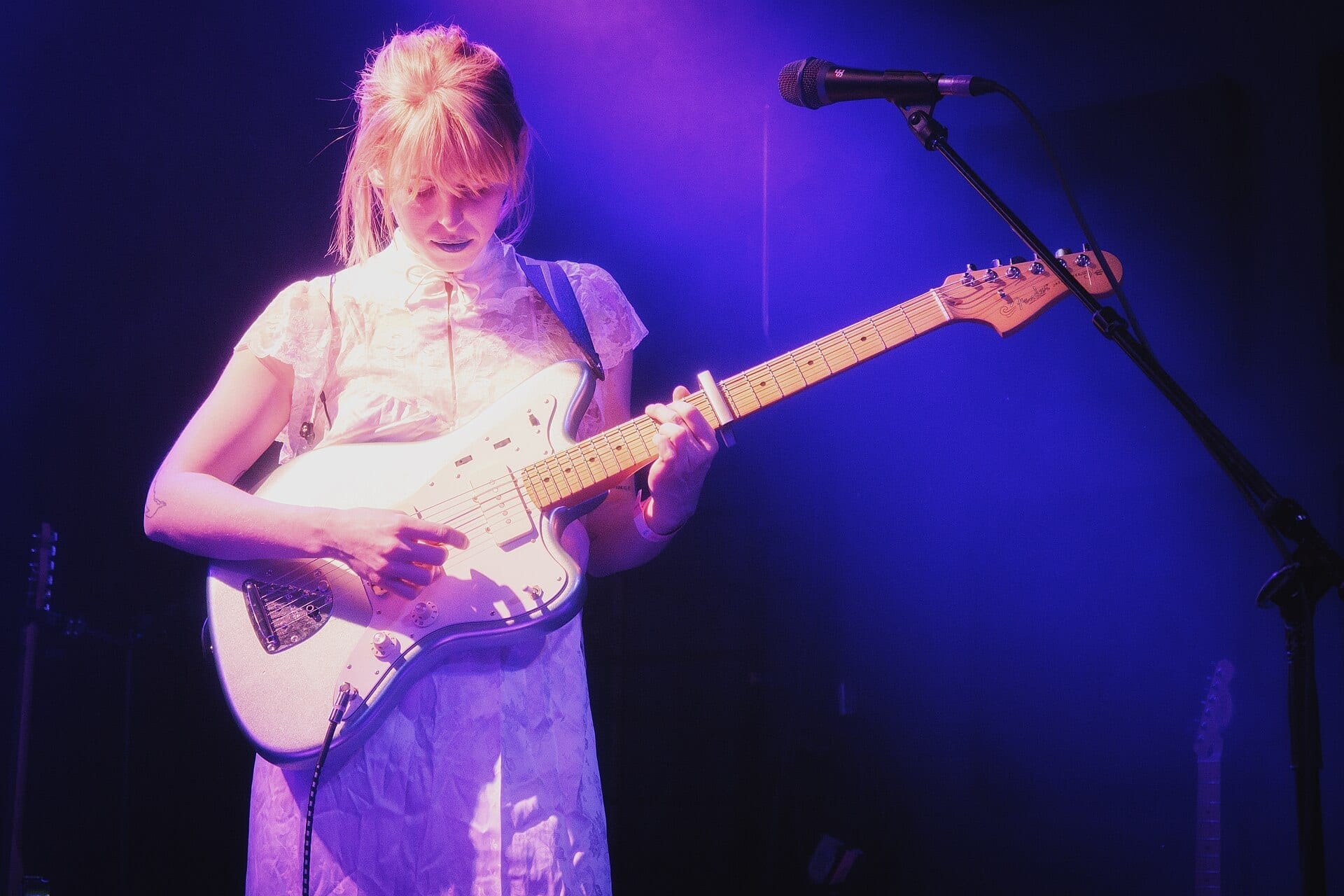 Fenne Lily plays a white electric guitar and sings into a microphone on a dimly lit stage bathed in purple light, her blonde hair pulled back with bangs falling across her forehead.