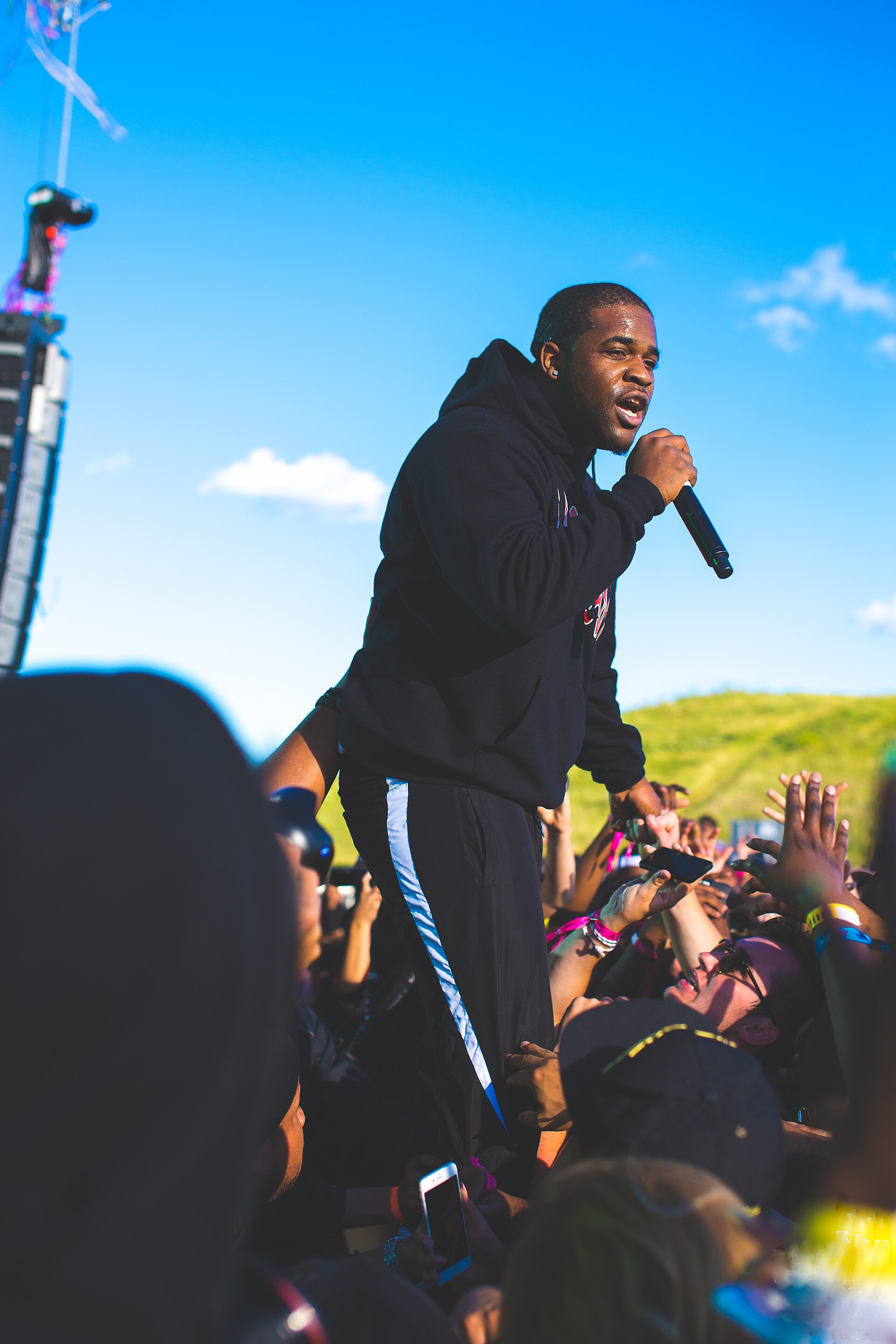 Ferg raps into a handheld microphone in a black hoodie and track pants, standing at the barrier under a blue sky with fans reaching up around him.