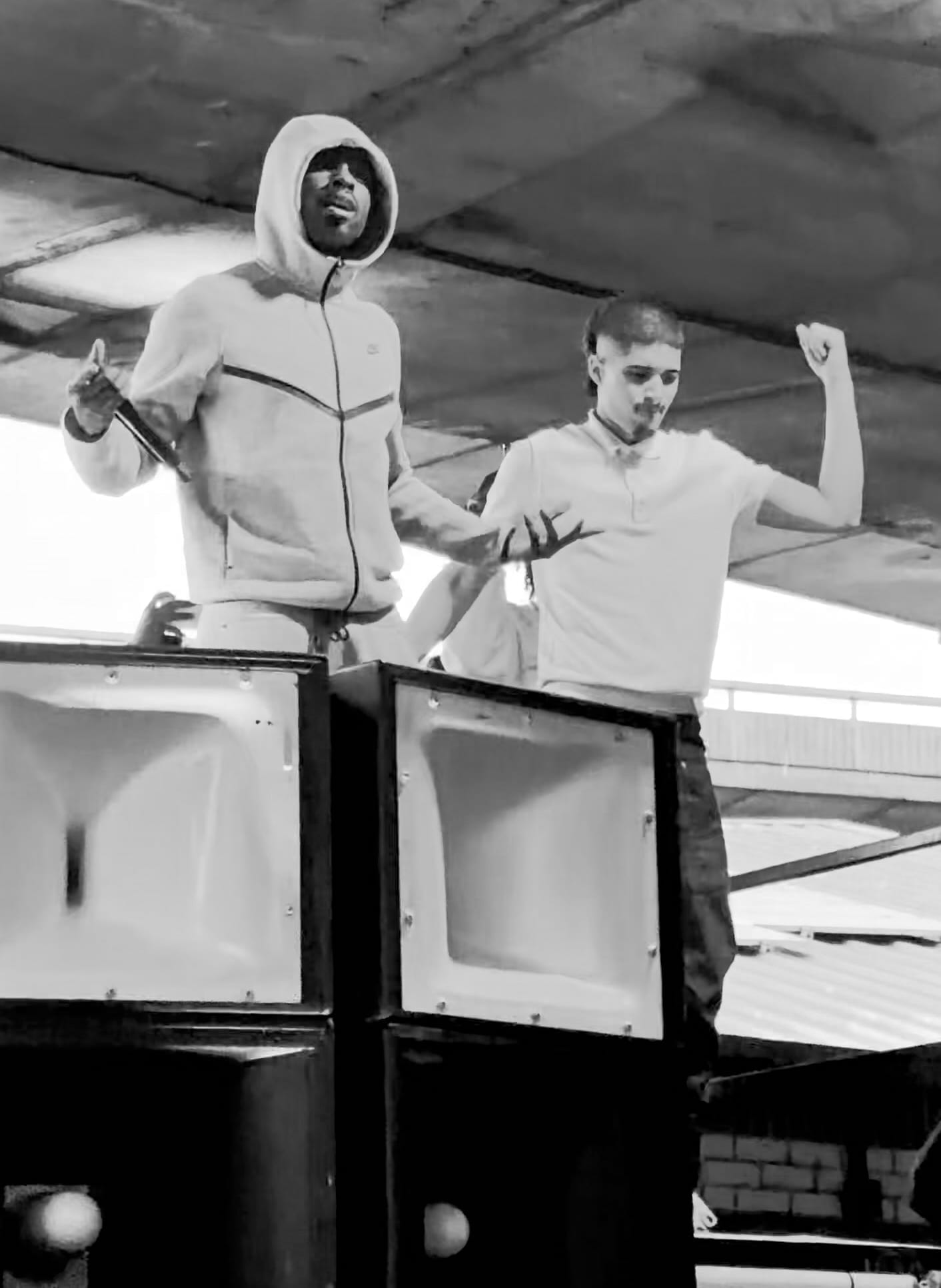 A grainy black-and-white photo of Fimiguerrero in a hooded jacket, holding a microphone, performing atop a stack of speakers under a car park roof alongside another rapper.