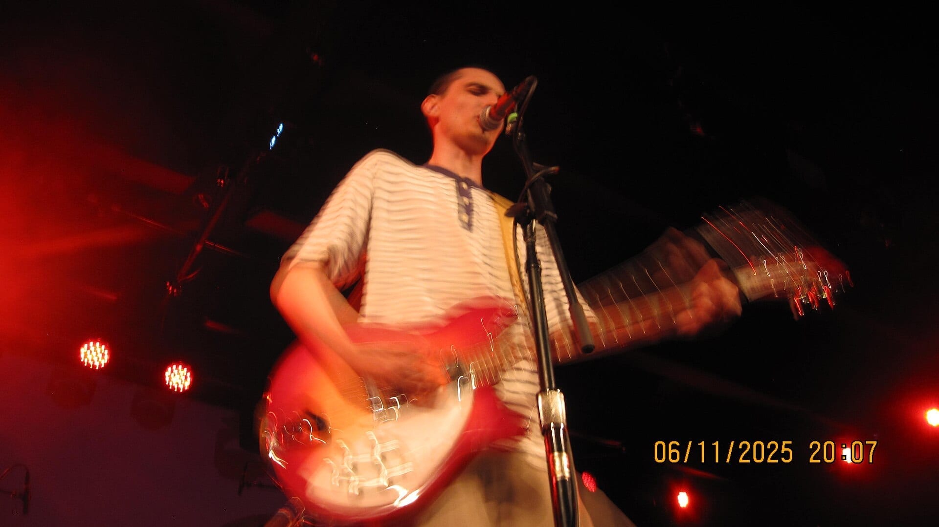 Finn Wolfhard sings into a microphone while playing a red electric guitar under red stage lights, captured mid-motion.